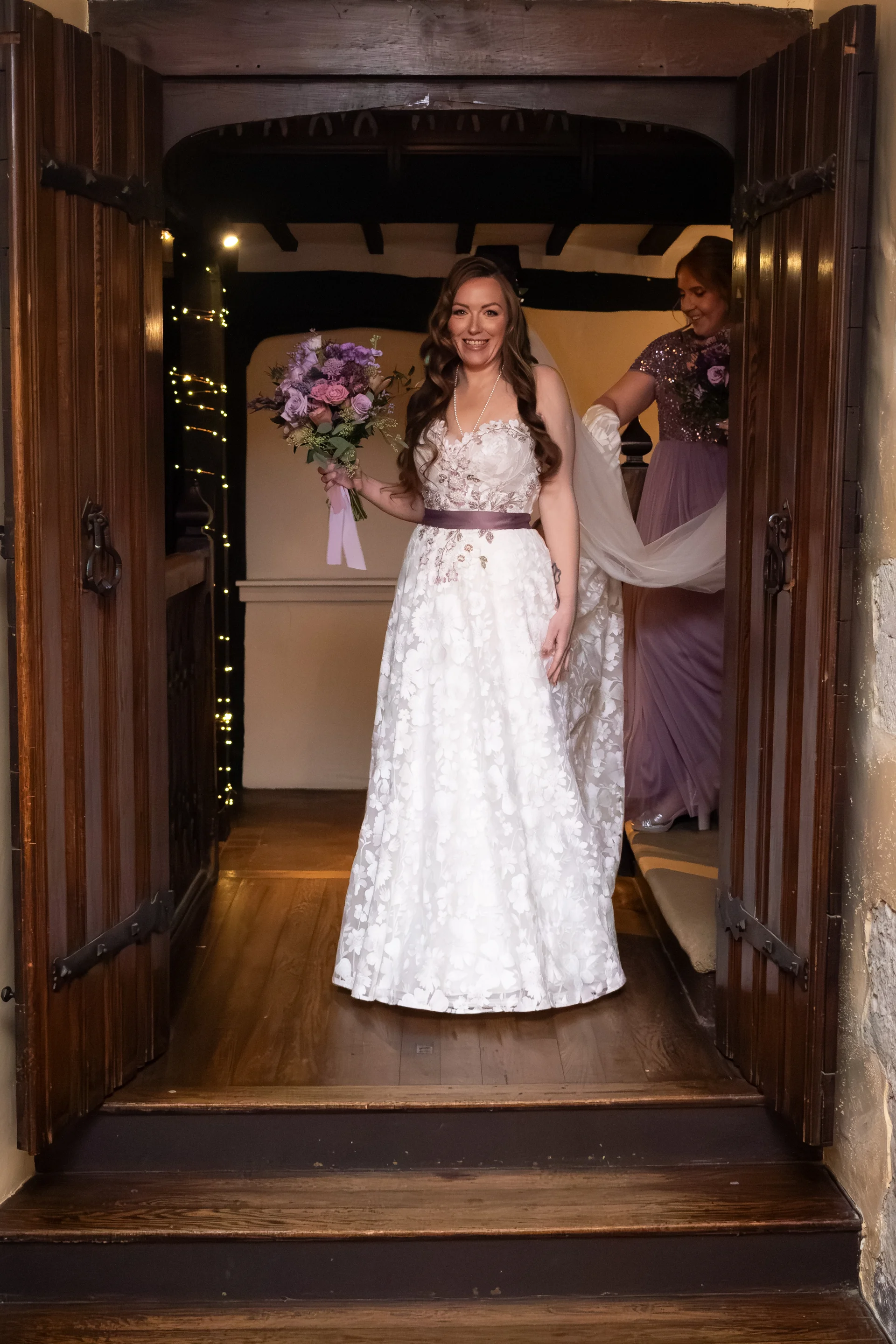 A smiling bride holding a purple floral bouquet stands framed in a wooden doorway with her bridesmaid helping adjust her veil inside.