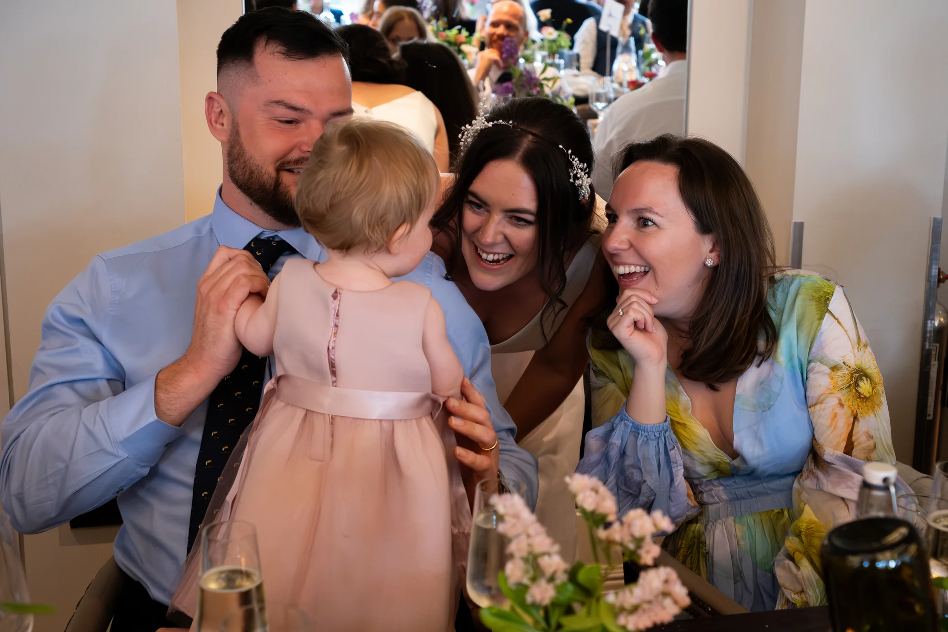 A joyful wedding guest and bride laugh delightedly as a young child in a pink dress is held up at the table during the reception.