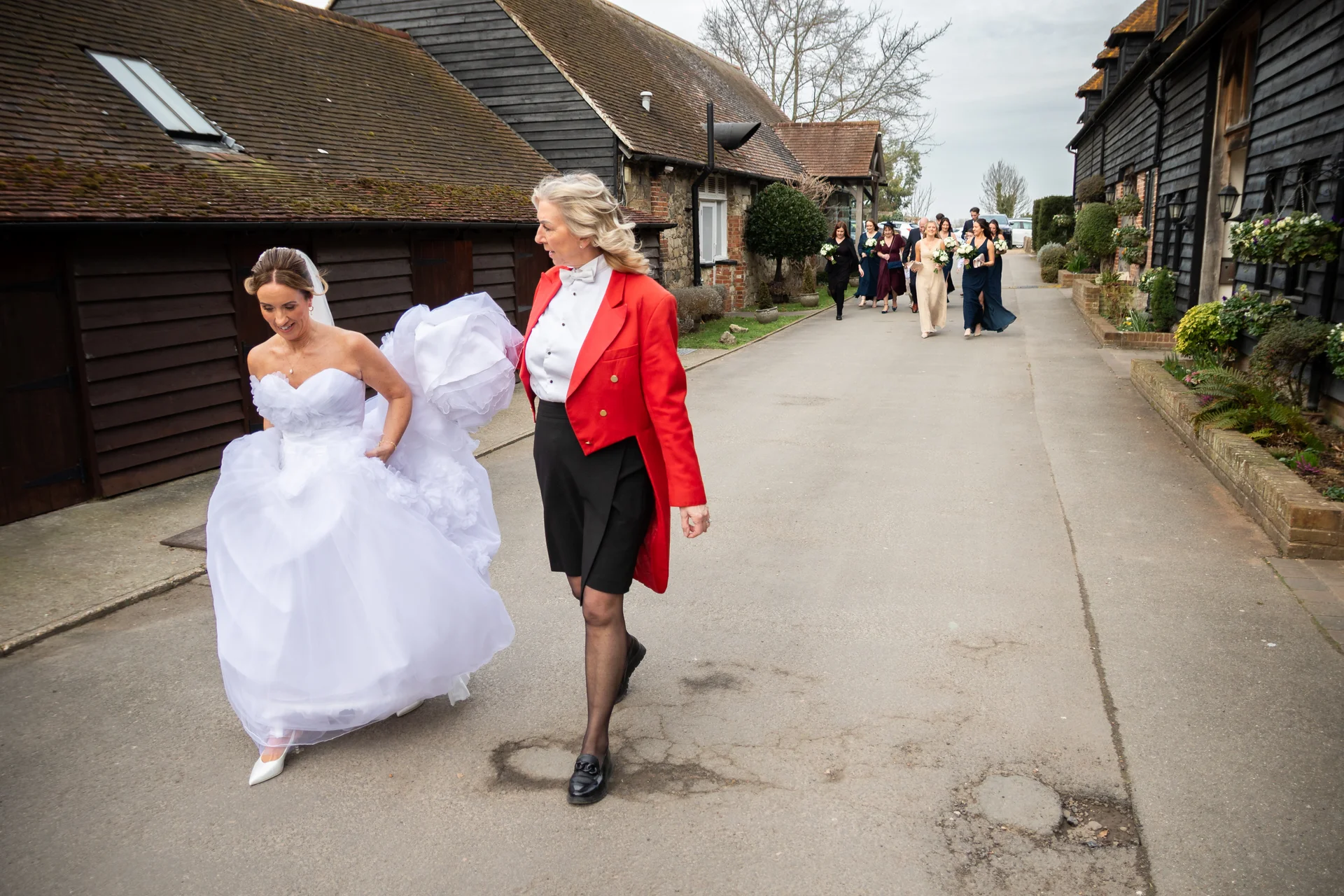A smiling bride in a voluminous strapless white gown and veil walks alongside a toastmaster in a red jacket outdoors between rustic timber-clad barns.
