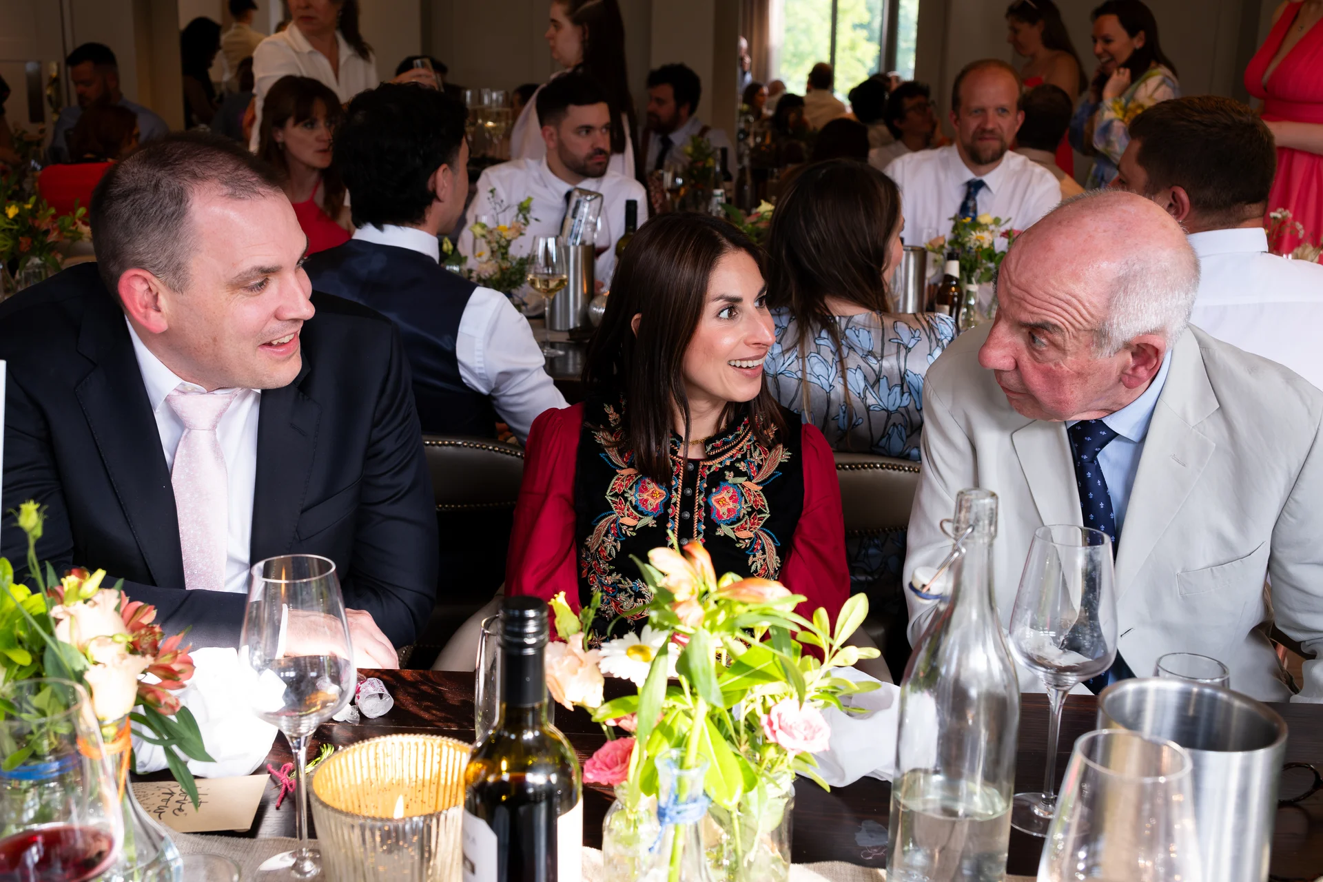 Wedding guests sit and converse animatedly around a long banquet table adorned with floral centrepieces and wine bottles during the wedding breakfast.