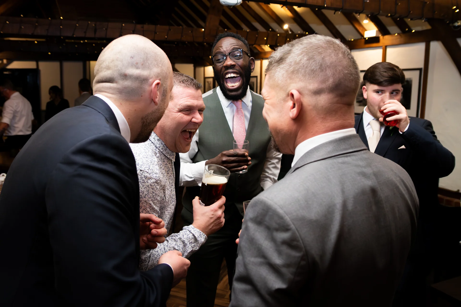 A group of groomsmen share a joyful laugh while holding drinks during a lively evening reception inside a rustic barn venue.