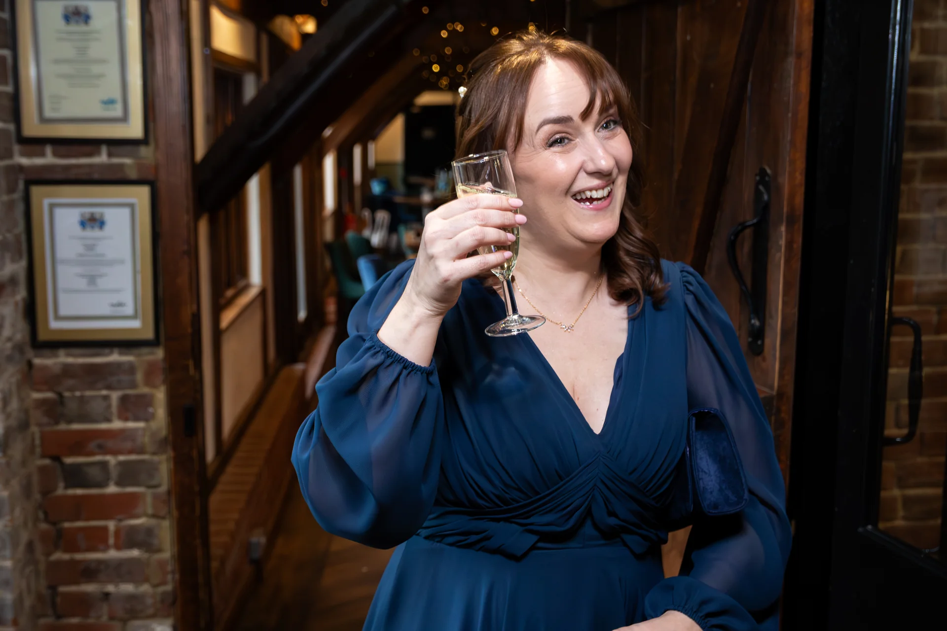 A smiling guest wearing a blue V-neck dress with sheer balloon sleeves holds up a glass of champagne during a wedding reception inside a rustic venue.