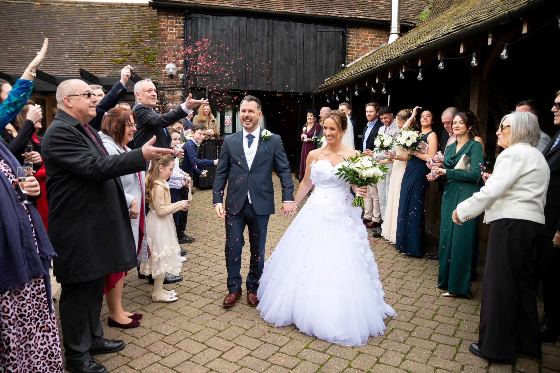 Newlyweds walk hand-in-hand through a cheering crowd of wedding guests who shower them with red confetti in a rustic barn courtyard.