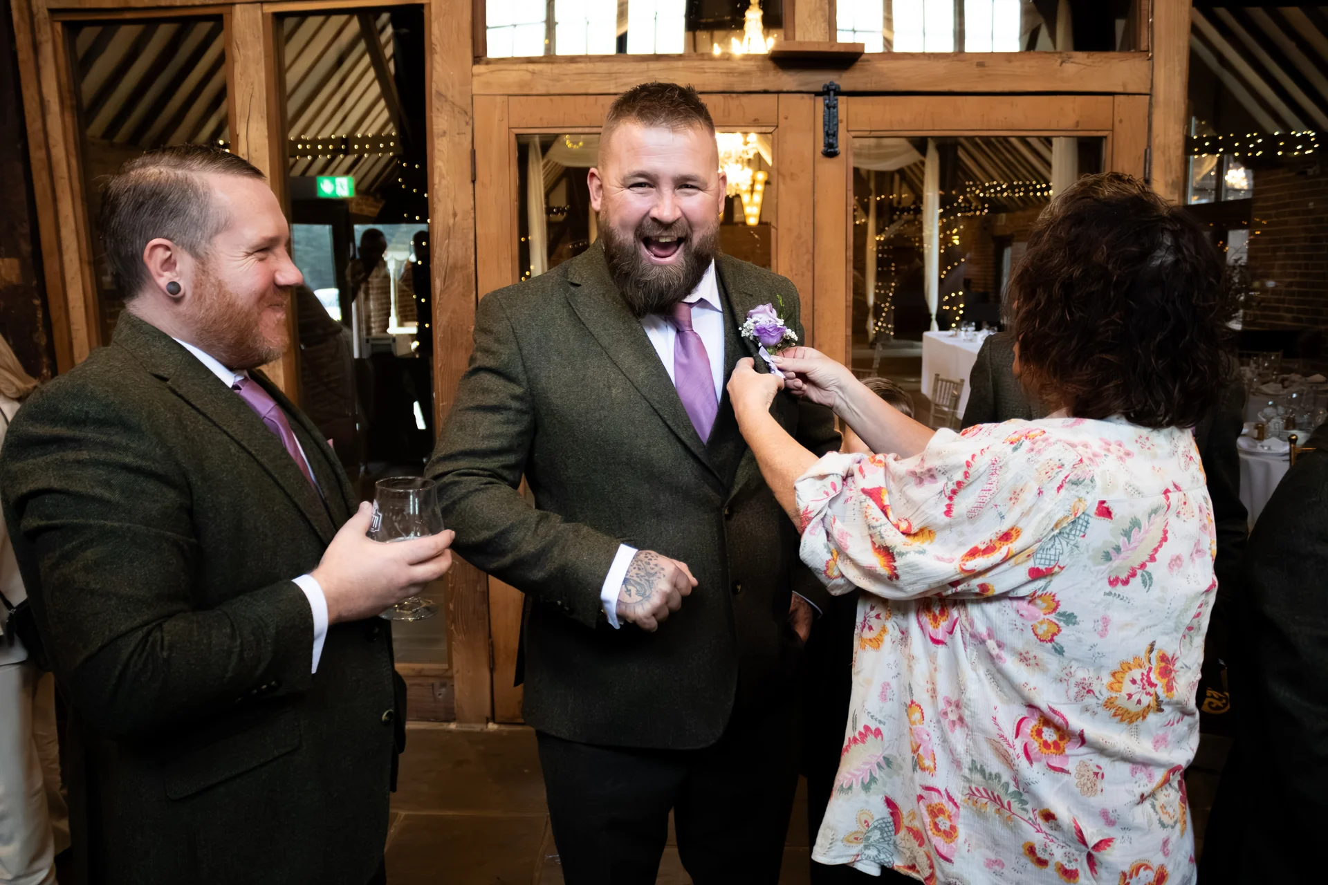 A joyful groom in an olive green tweed suit laughs as a guest pins a purple boutonniere to his lapel inside a rustic wooden barn venue.