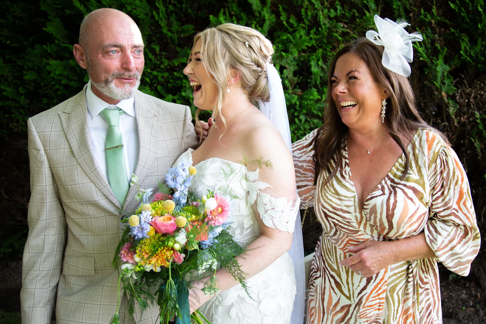 A joyful bride holding a colourful bouquet shares a hearty laugh with her father in a tweed suit and a smiling guest wearing a fascinator.