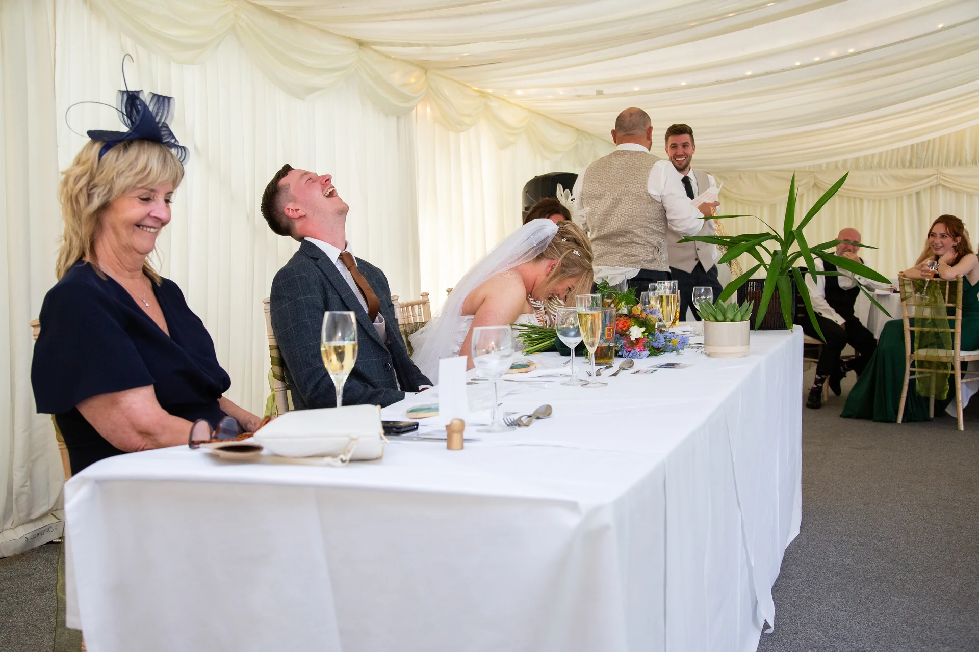 A groom throws his head back in laughter during the wedding speeches at a marquee reception, while family and guests seated at the top table smile and laugh along.