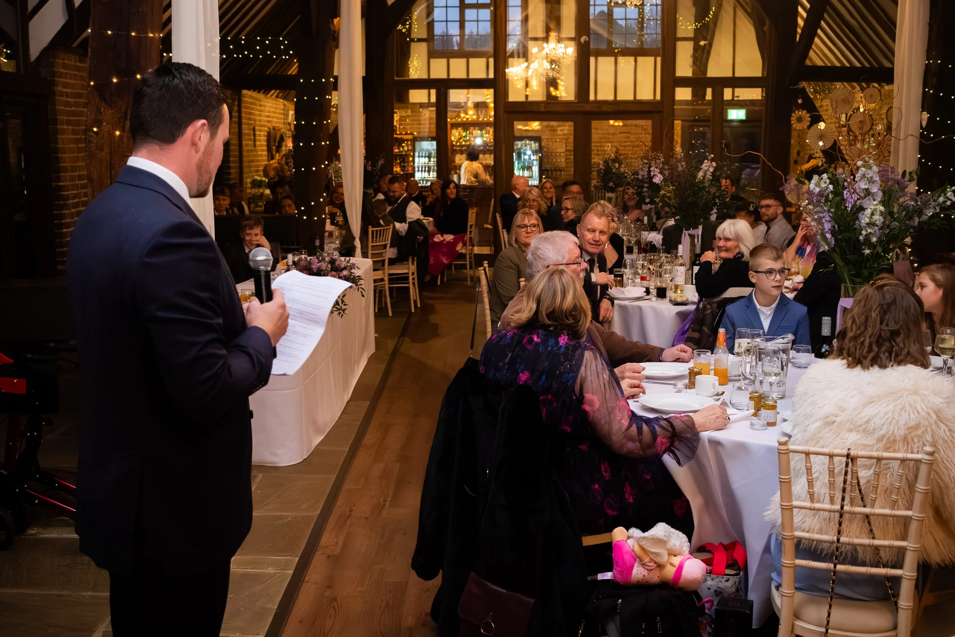 A groom in a dark suit stands delivering a speech holding notes and a microphone in front of seated wedding guests inside a rustic barn venue illuminated by warm fairy lights.