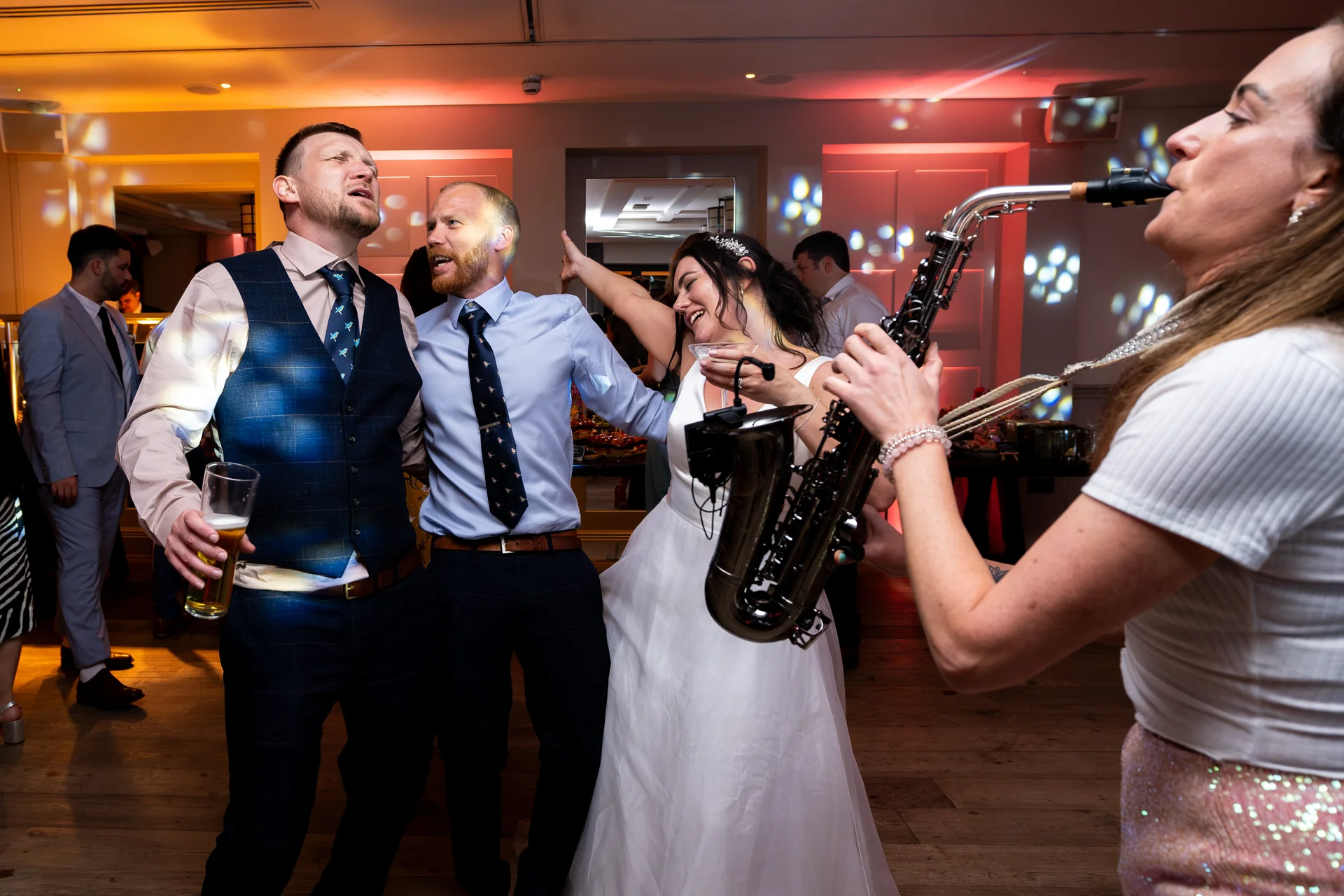 A joyous bride and groom dancing energetically on a packed dance floor alongside guests while a saxophonist performs.