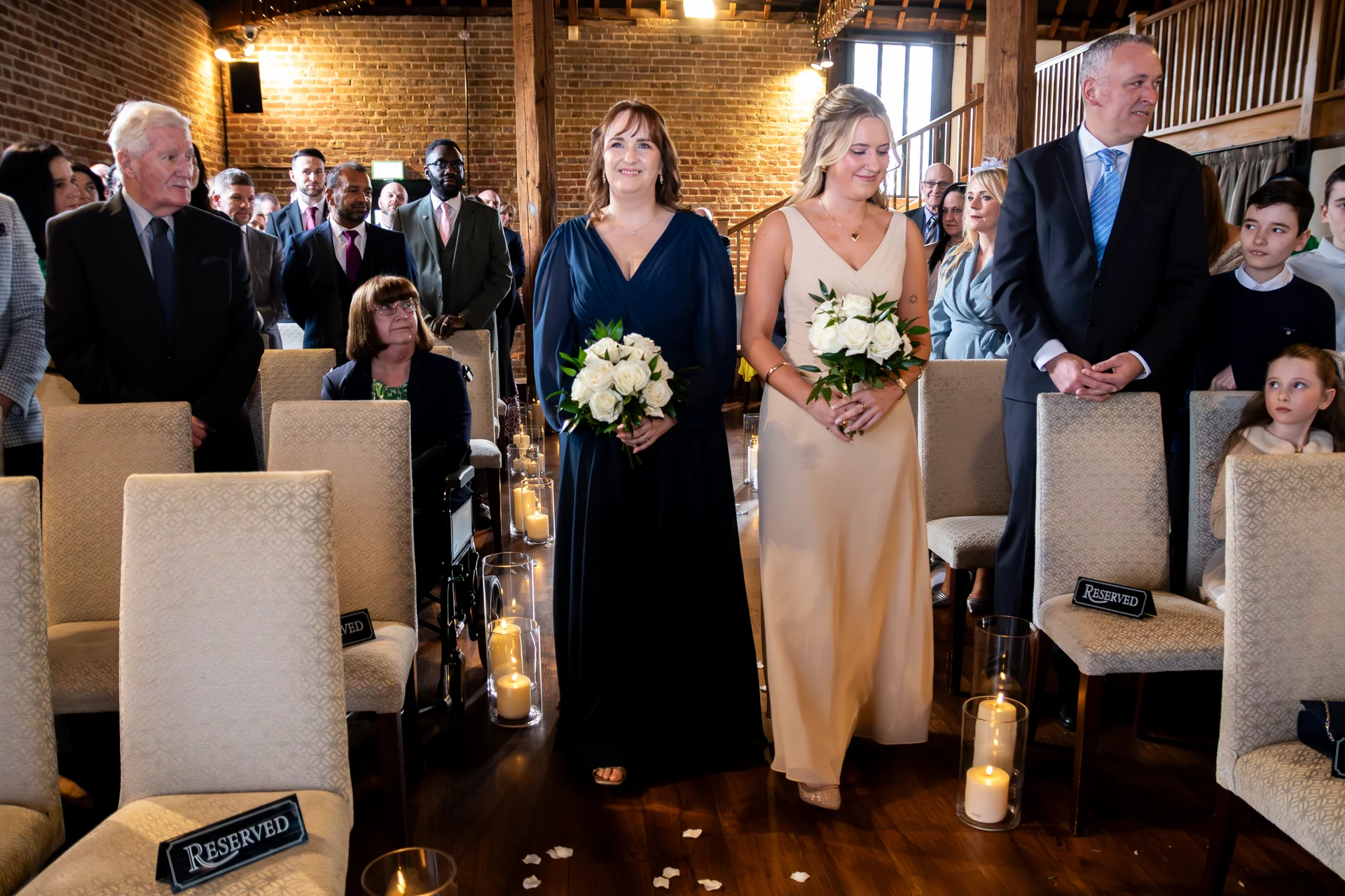 Two bridesmaids hold white rose bouquets while walking down the aisle during a ceremony inside a rustic brick barn venue adorned with floor candles.