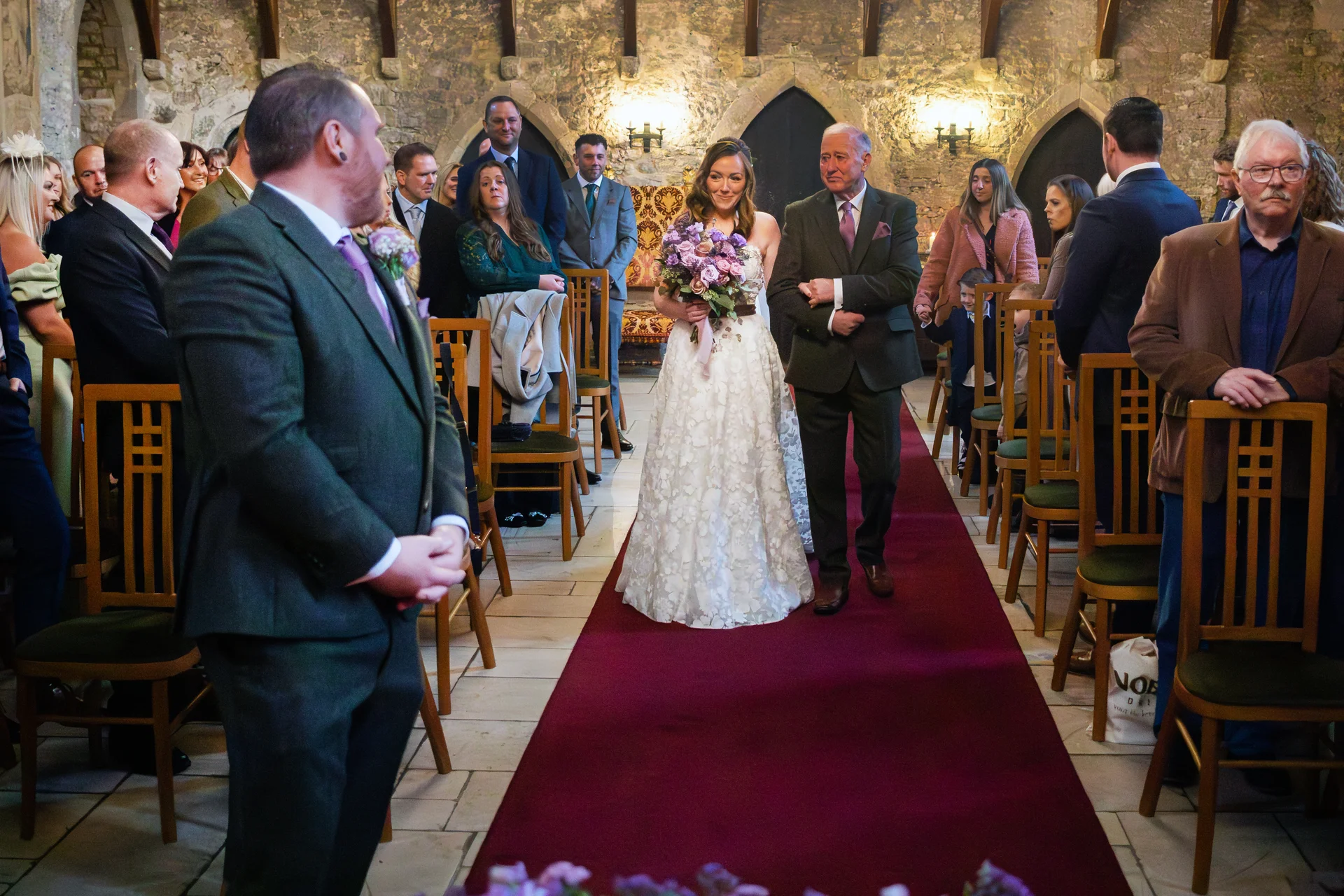 A smiling bride walks down a red-carpeted aisle inside a historic castle venue, linked arm-in-arm with her father while holding a bouquet of purple flowers, as the groom watches from the front.