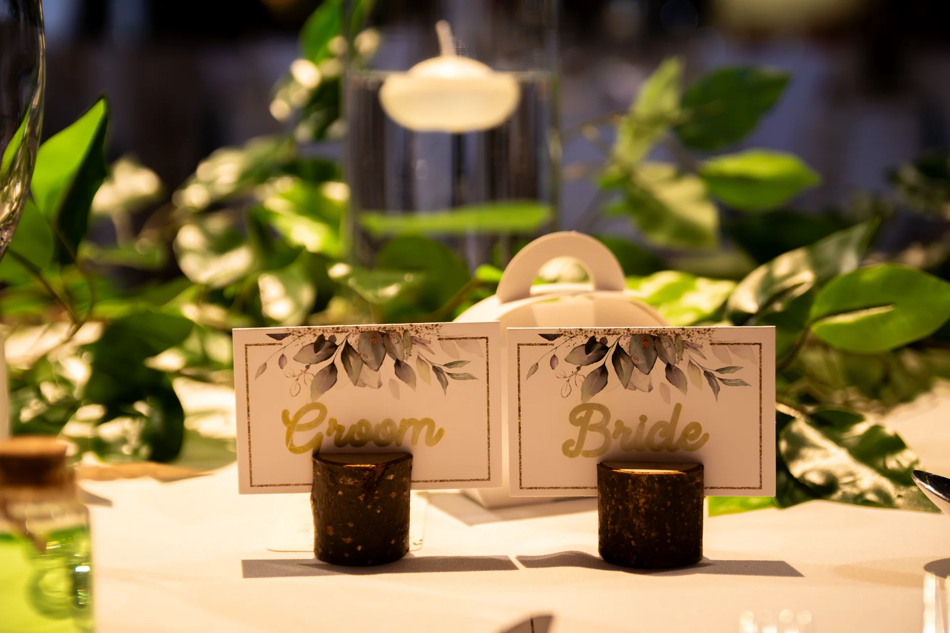 Close-up of wooden place card holders displaying "Groom" and "Bride" cards on a wedding tablescape adorned with lush green foliage and a floating candle.