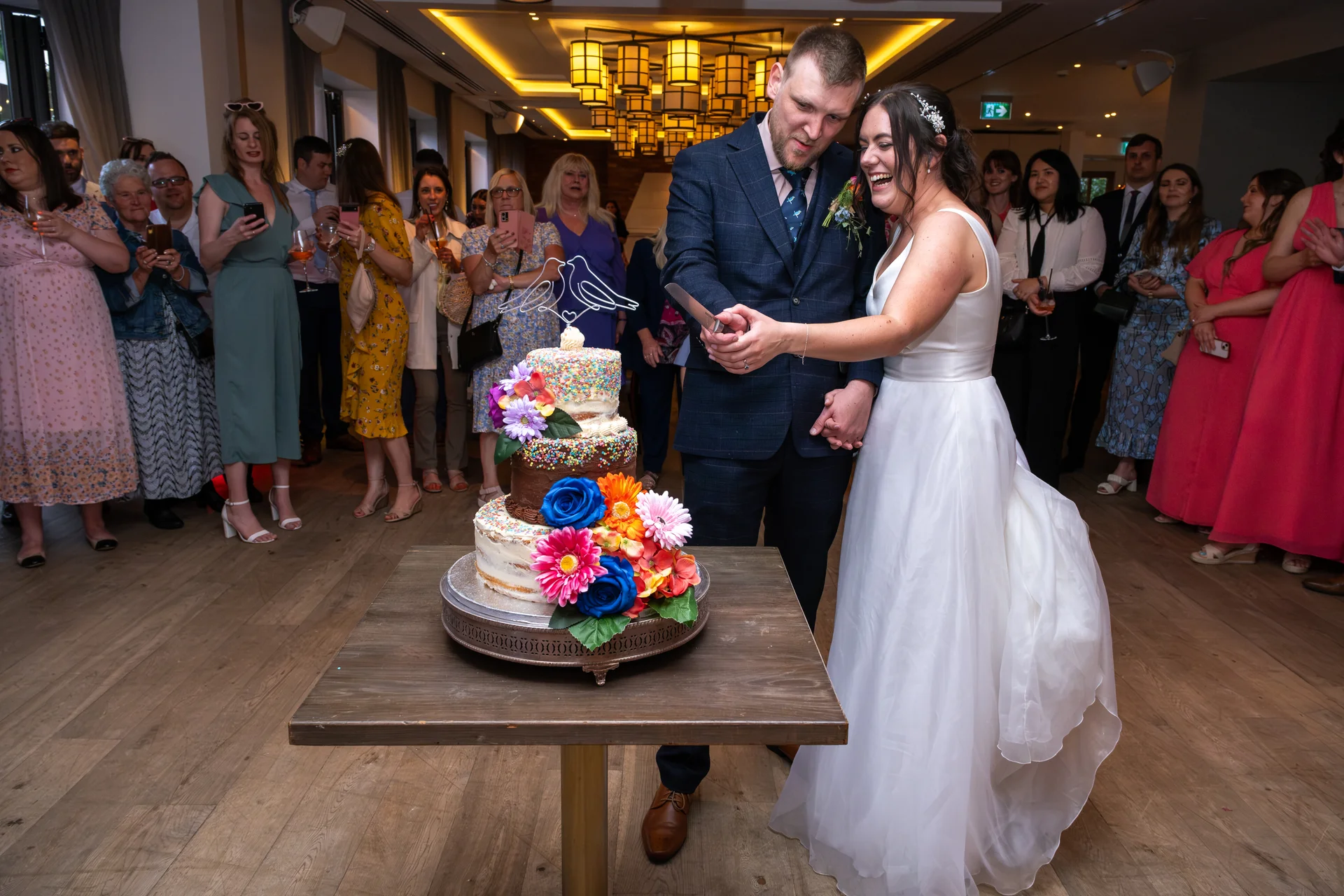 The bride and groom laugh gleefully while cutting a multi-tiered sprinkle cake decorated with vibrant floral arrangements, surrounded by cheering wedding guests inside a modern venue.
