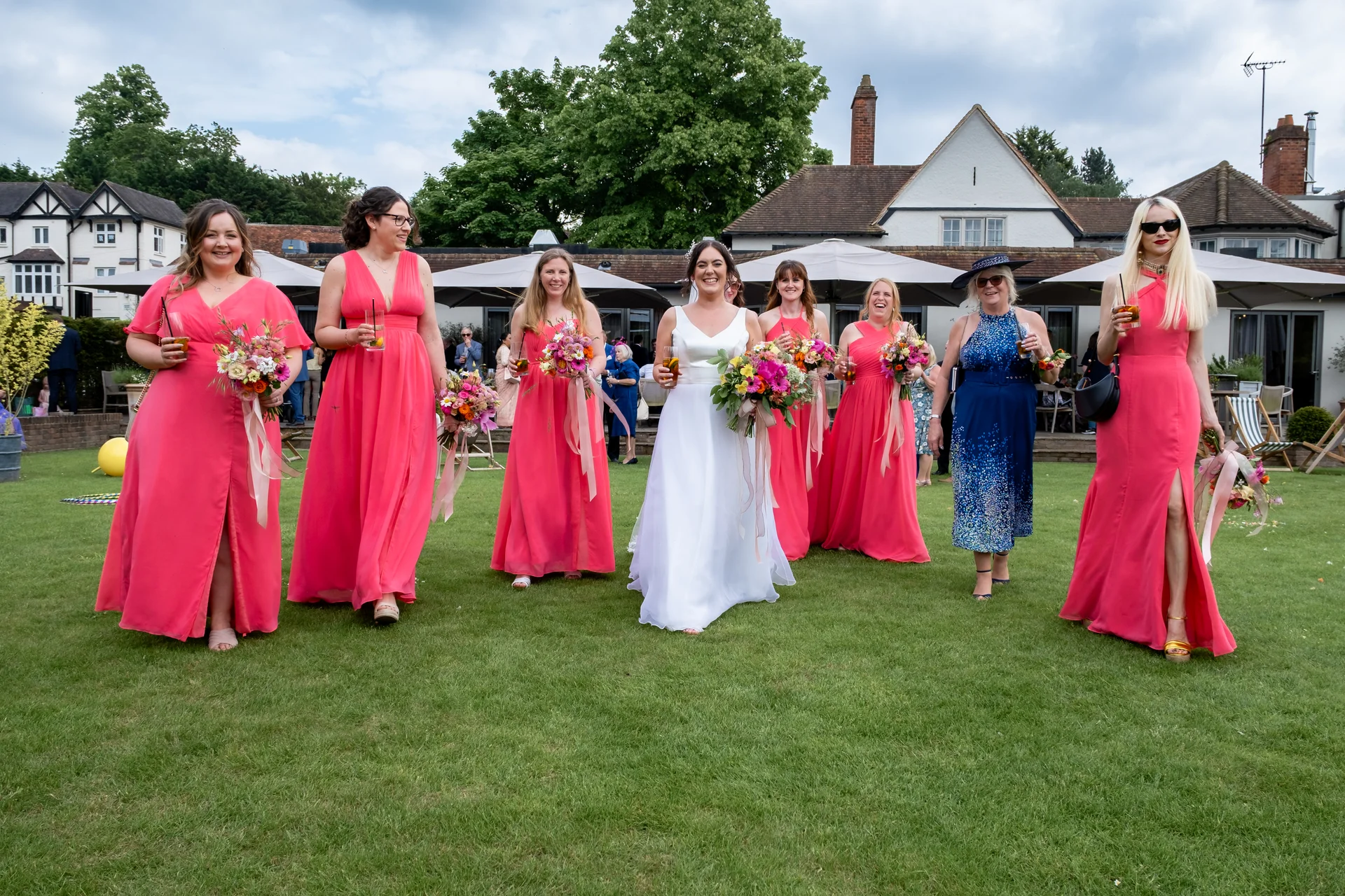 A smiling bride and her bridesmaids in vibrant pink dresses walk across a lush green lawn during a country house wedding drinks reception.