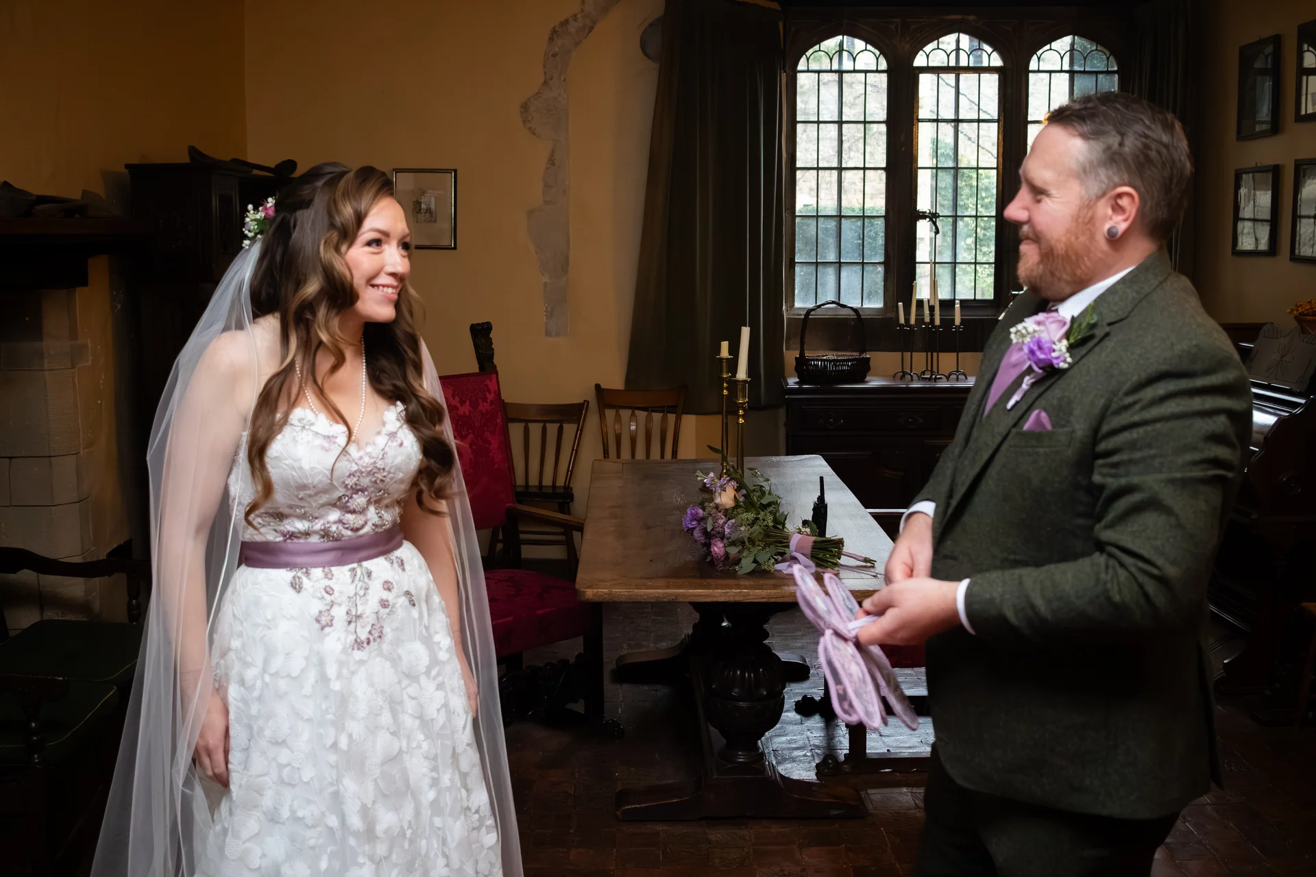 A smiling bride in a floral-detailed white gown with a purple sash shares a tender first look with her groom, who wears a green tweed suit in a historic manor house.