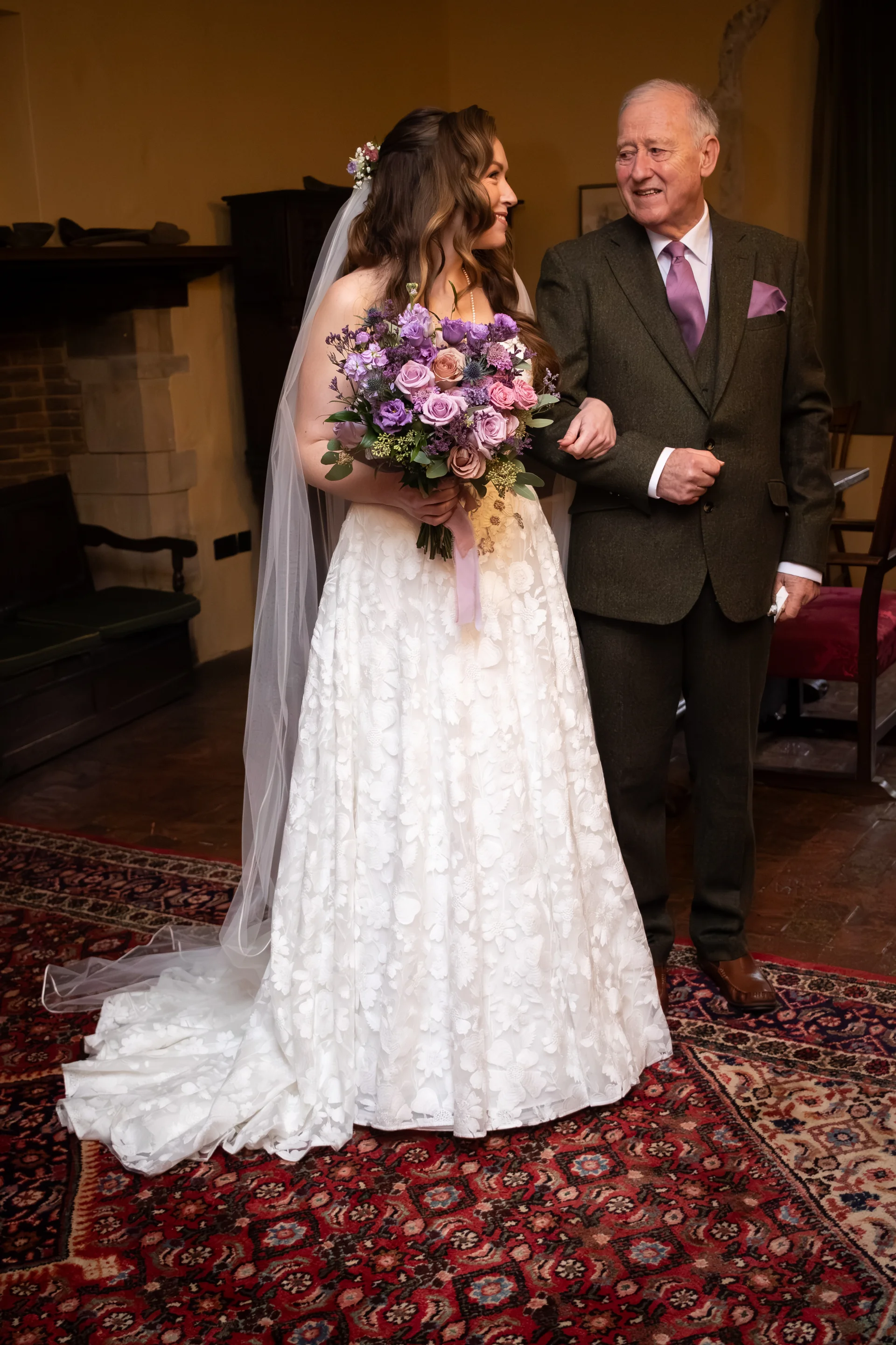 A smiling bride holding a purple floral bouquet stands arm in arm with her father, who is wearing a green tweed suit inside a rustic venue.