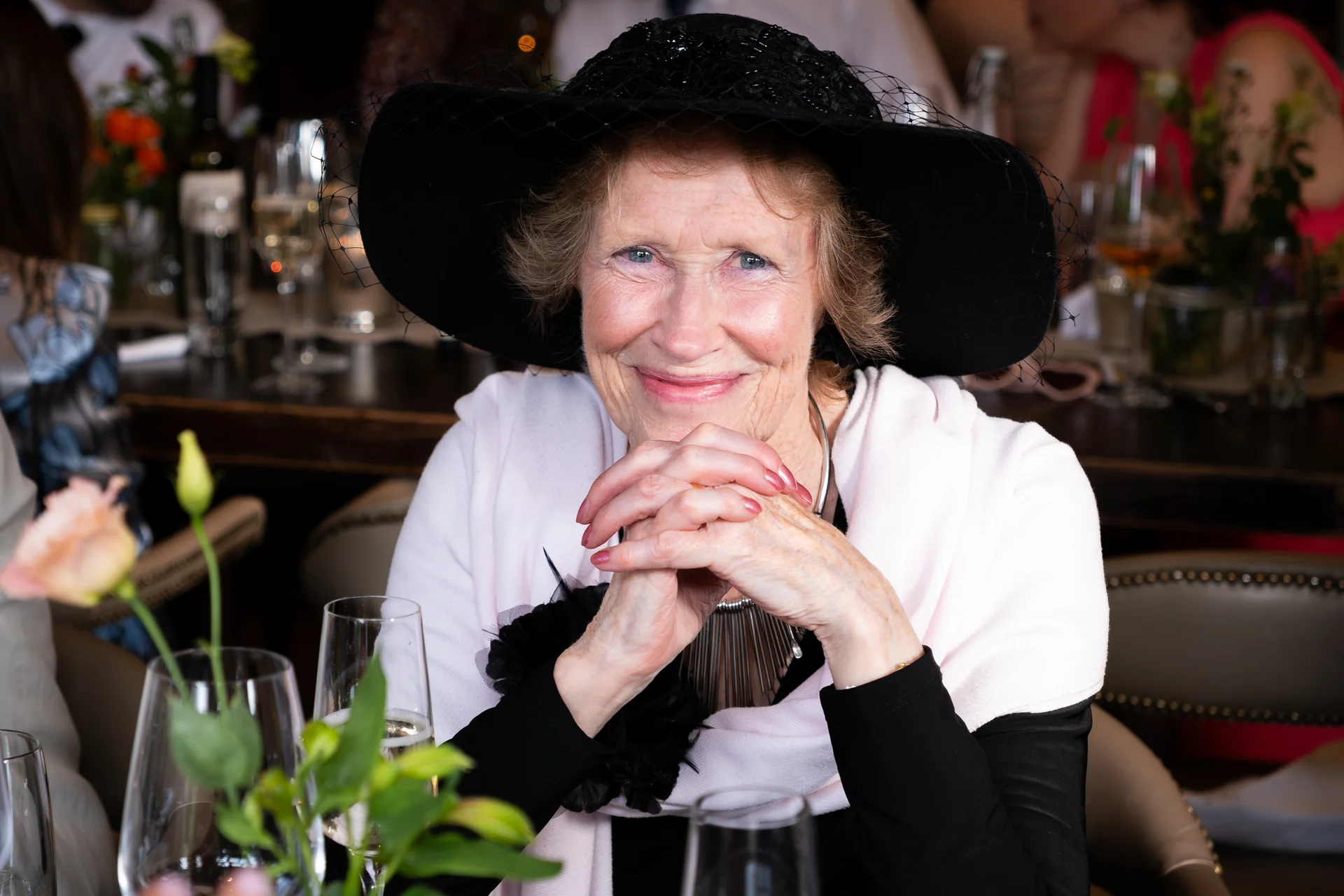 An elderly female guest wearing a wide-brimmed black hat and pink wrap smiles warmly with her hands clasped at a wedding reception table.