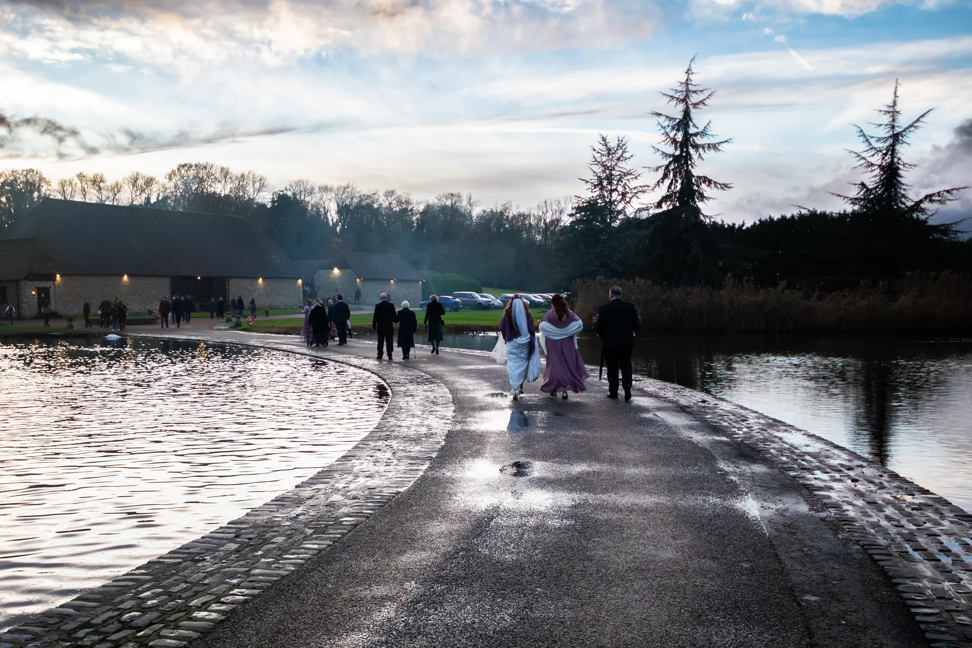 Wedding guests walk along a curved stone path beside a calm lake towards a rustic barn venue at dusk under a dramatic sky.