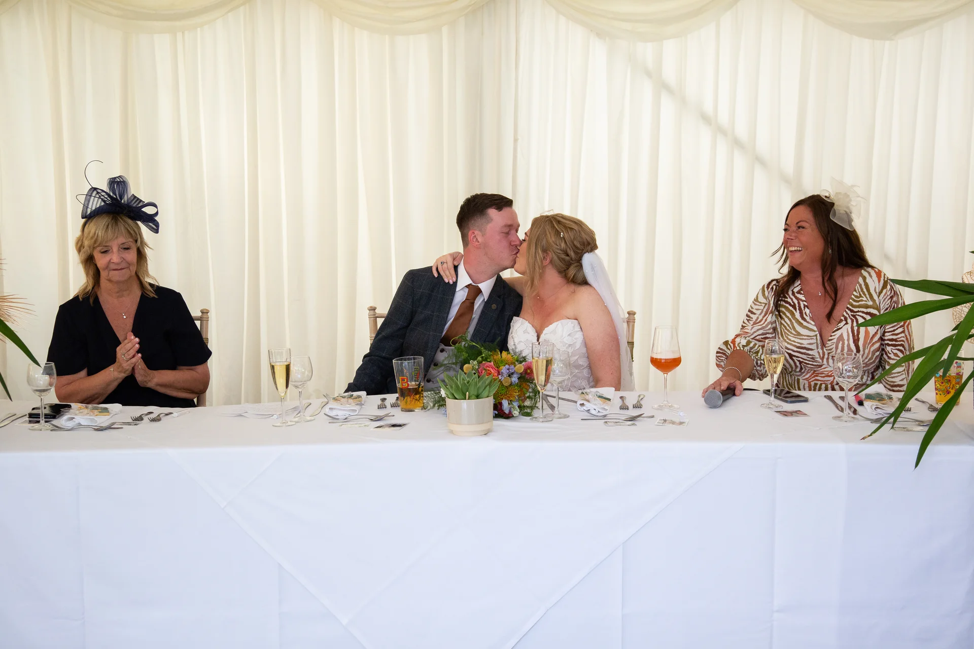 Newlyweds share a kiss at the top table during a marquee wedding breakfast while family members look on and smile.