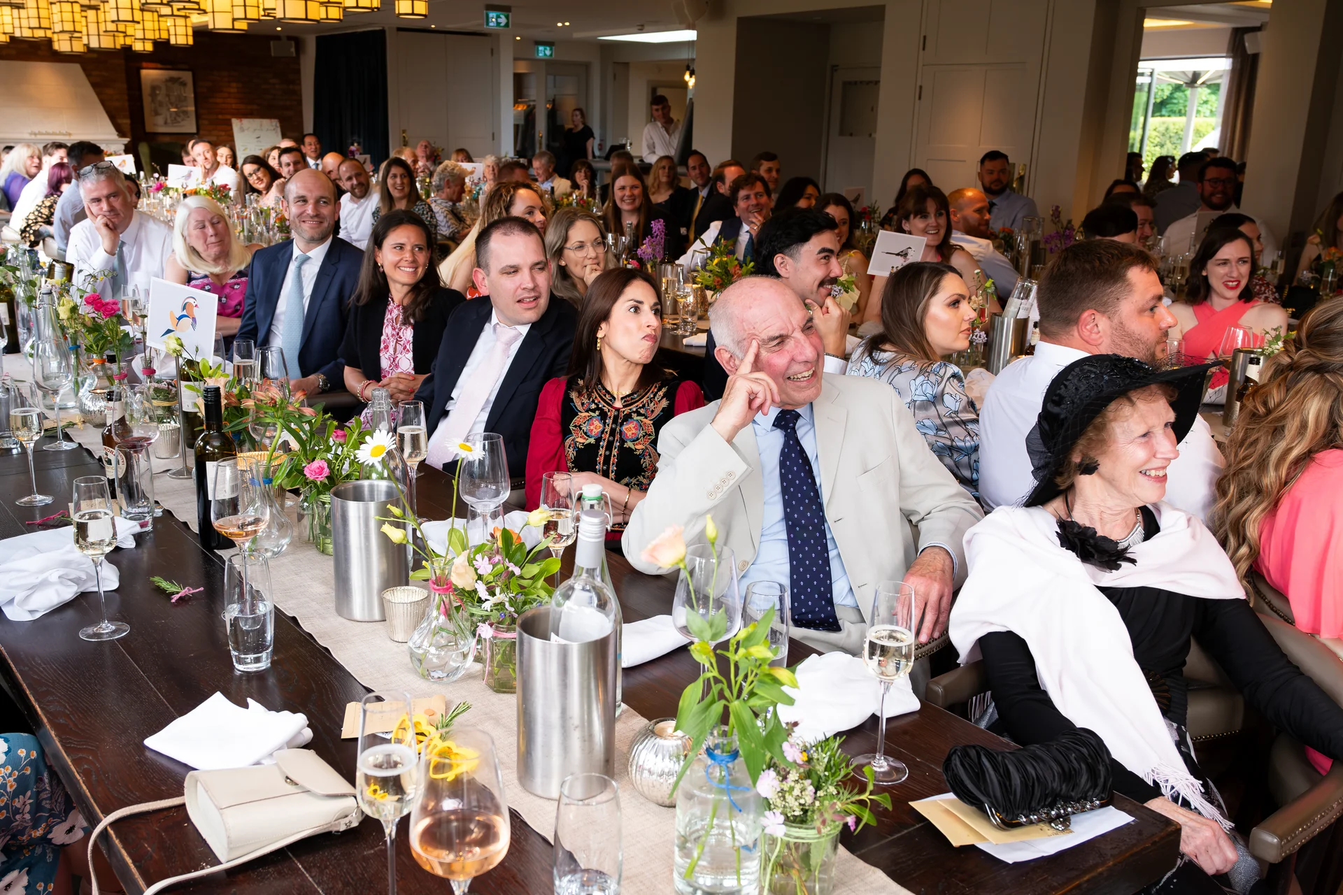 Wedding guests laugh heartily at the speeches during the wedding breakfast, seated along a long banquet table decorated with candles and floral centrepieces.