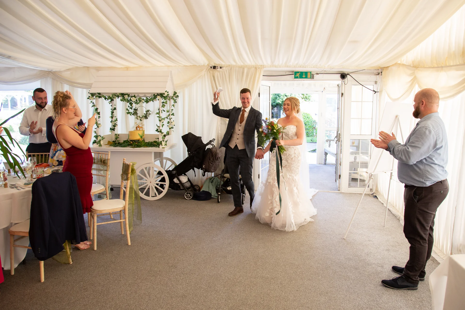 The joyful newlyweds enter their marquee wedding reception hand in hand, greeted by cheering guests and applause.