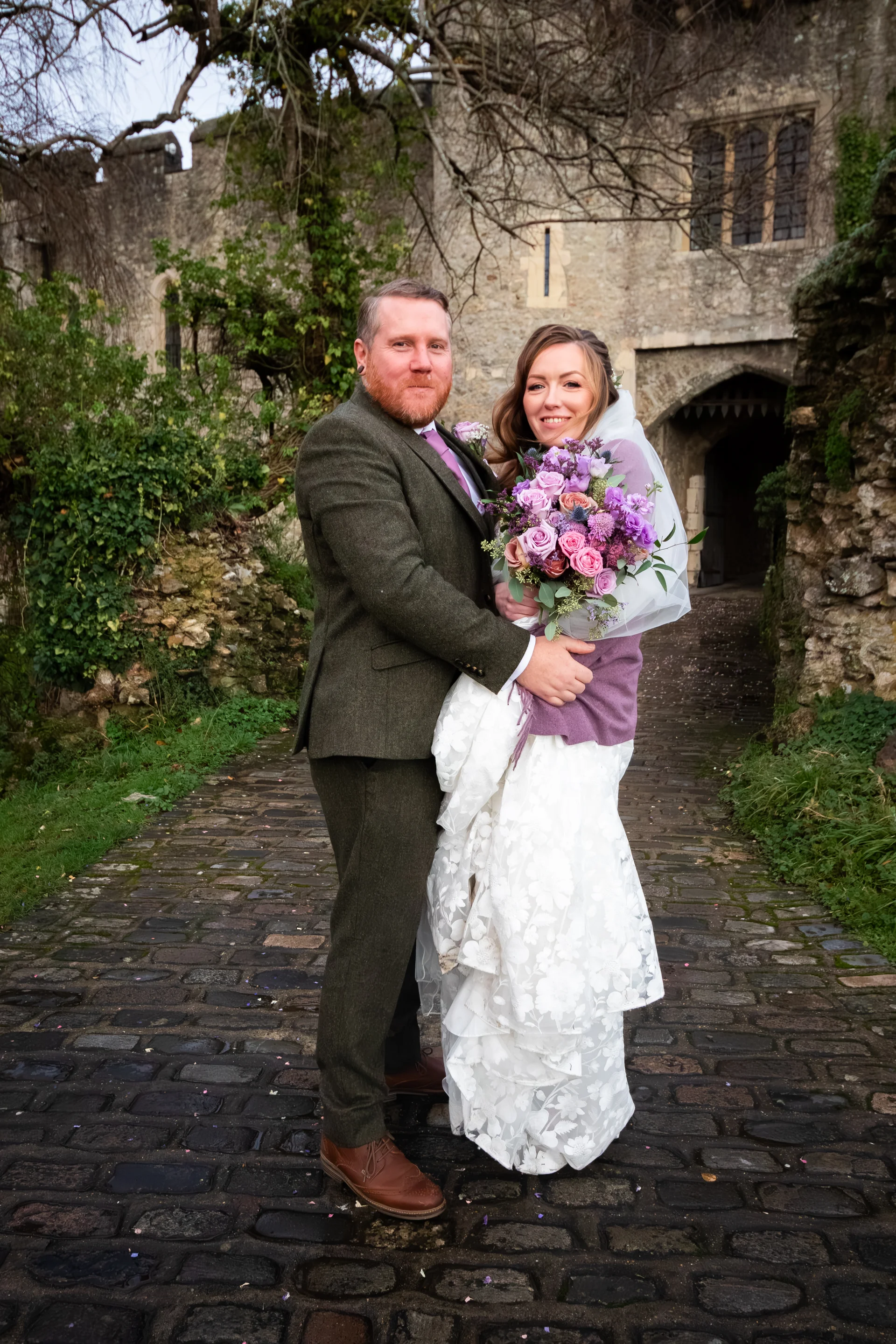 A smiling bride and groom pose together on a cobbled path outside an ancient stone castle, with the bride holding a colourful hand-tied bouquet of purple and pink flowers.