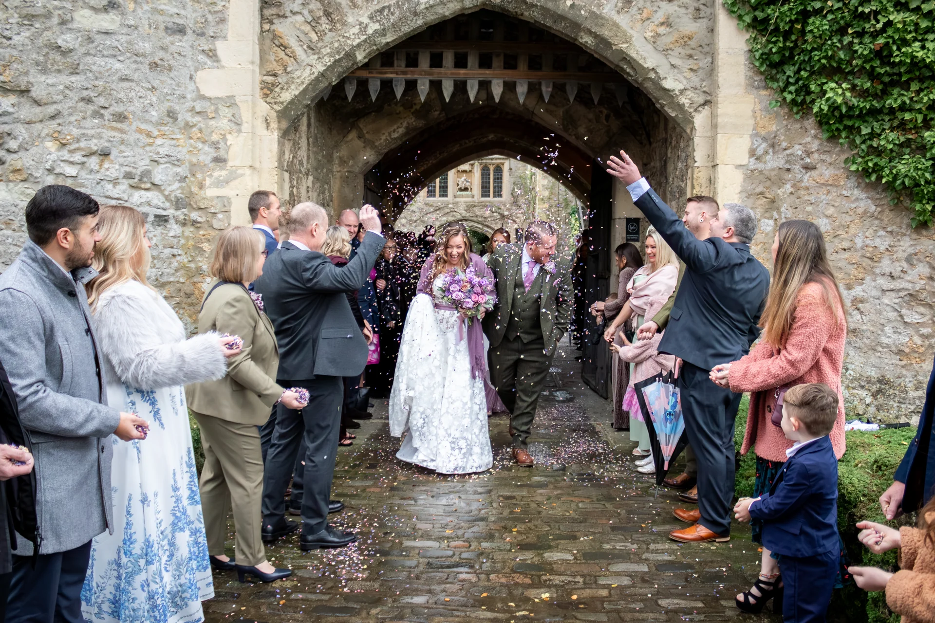 The bride and groom laugh joyfully as they walk through a historic stone archway while wedding guests shower them with colourful confetti.