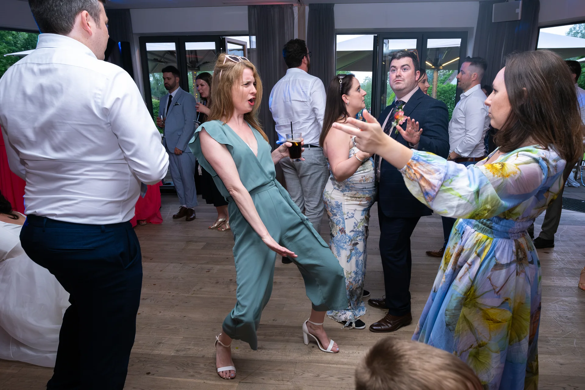 Wedding guests enjoy dancing on the wooden dance floor, with one woman in a sage green jumpsuit striking a playful pose while holding a drink.