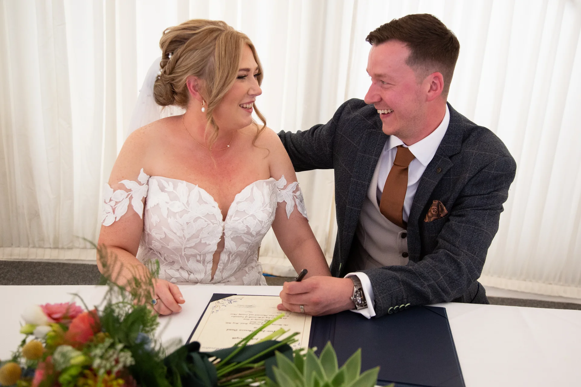 A joyful bride and groom smile warmly at each other while signing the marriage register during their wedding ceremony.