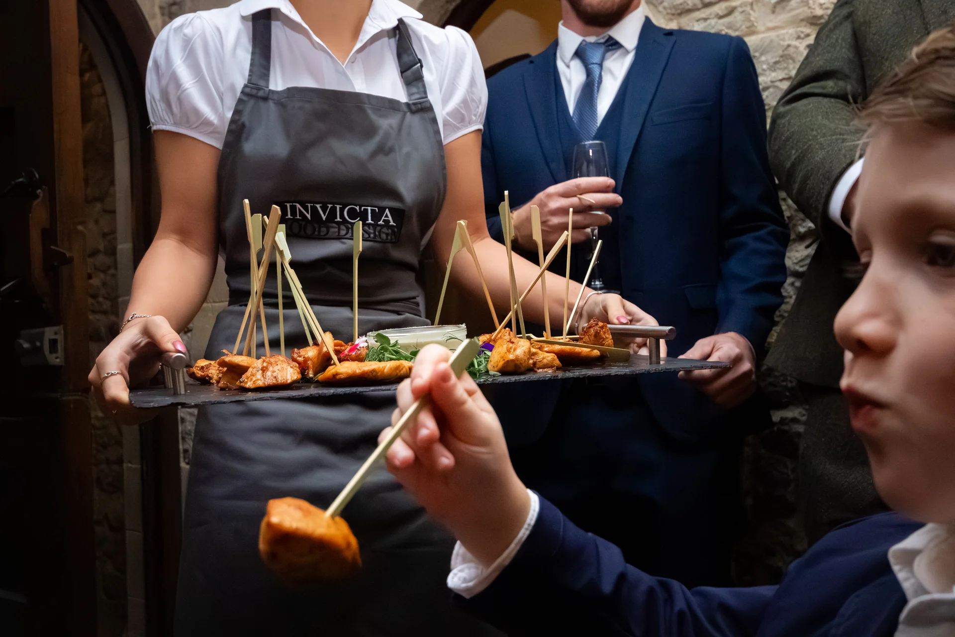 A young wedding guest in a blue suit uses a bamboo skewer to pick a canape from a slate tray held by a catering staff member.