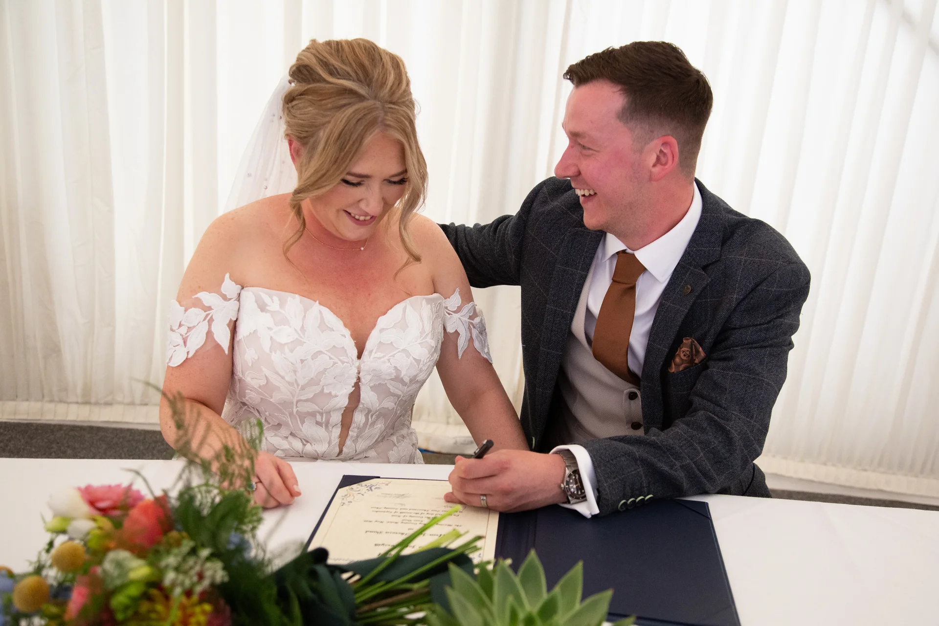 The bride and groom share a joyful laugh while signing the marriage register during their civil ceremony, seated at a white table decorated with a floral arrangement.