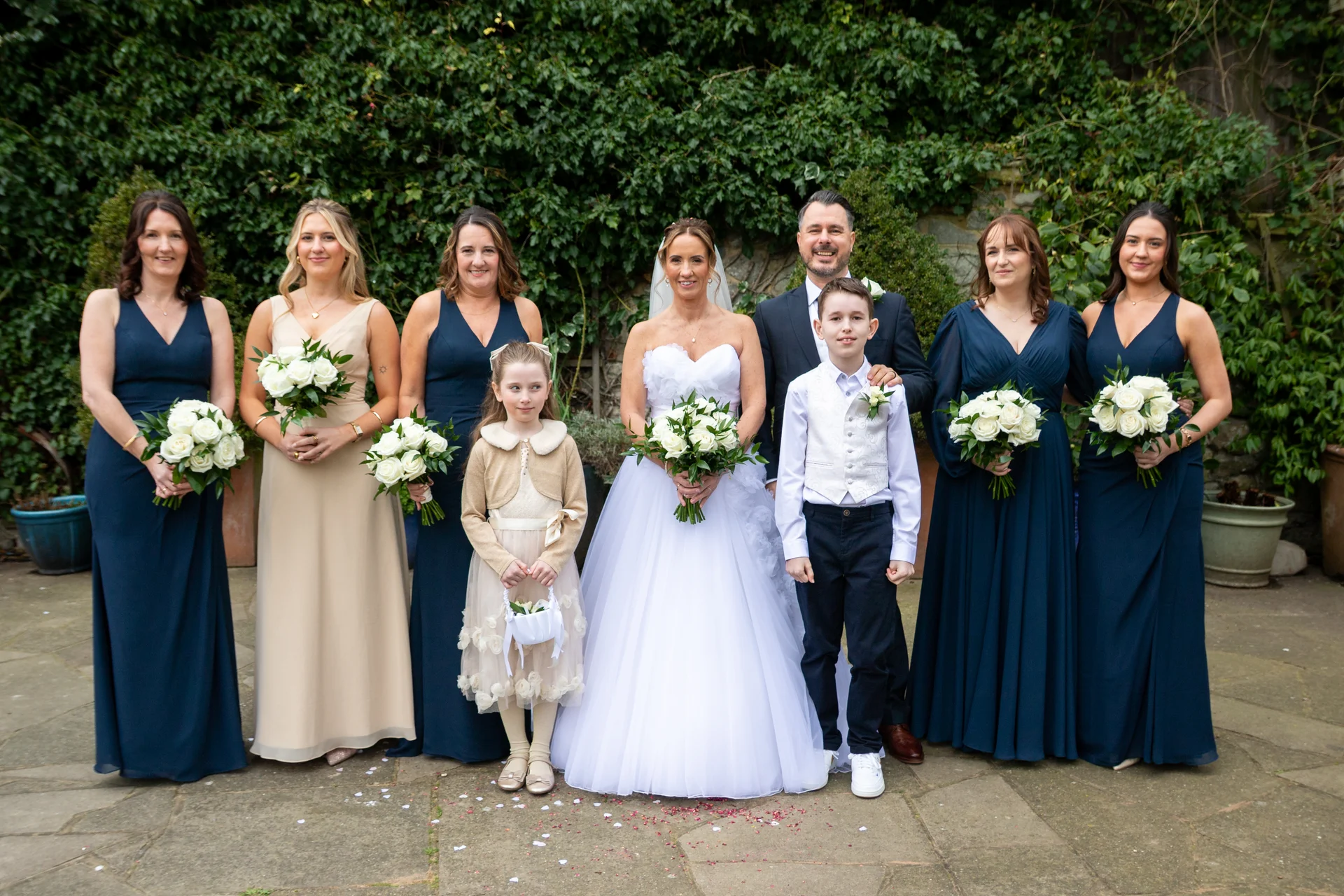 A bridal party comprising a bride in a strapless white gown, groom in a dark suit, bridesmaids in navy and champagne dresses, and a flower girl and page boy pose together outdoors in front of a stone wall covered in green ivy.