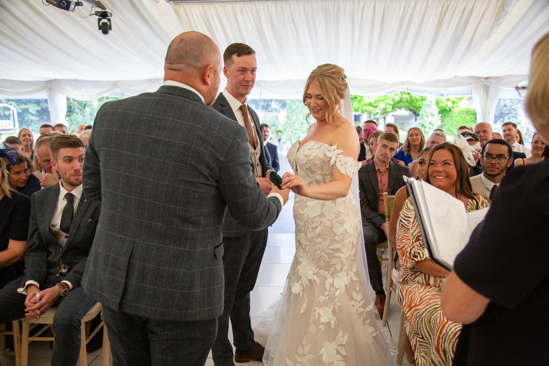 The bride and groom exchange rings during their wedding ceremony inside a marquee draped with white fabric, surrounded by seated guests.