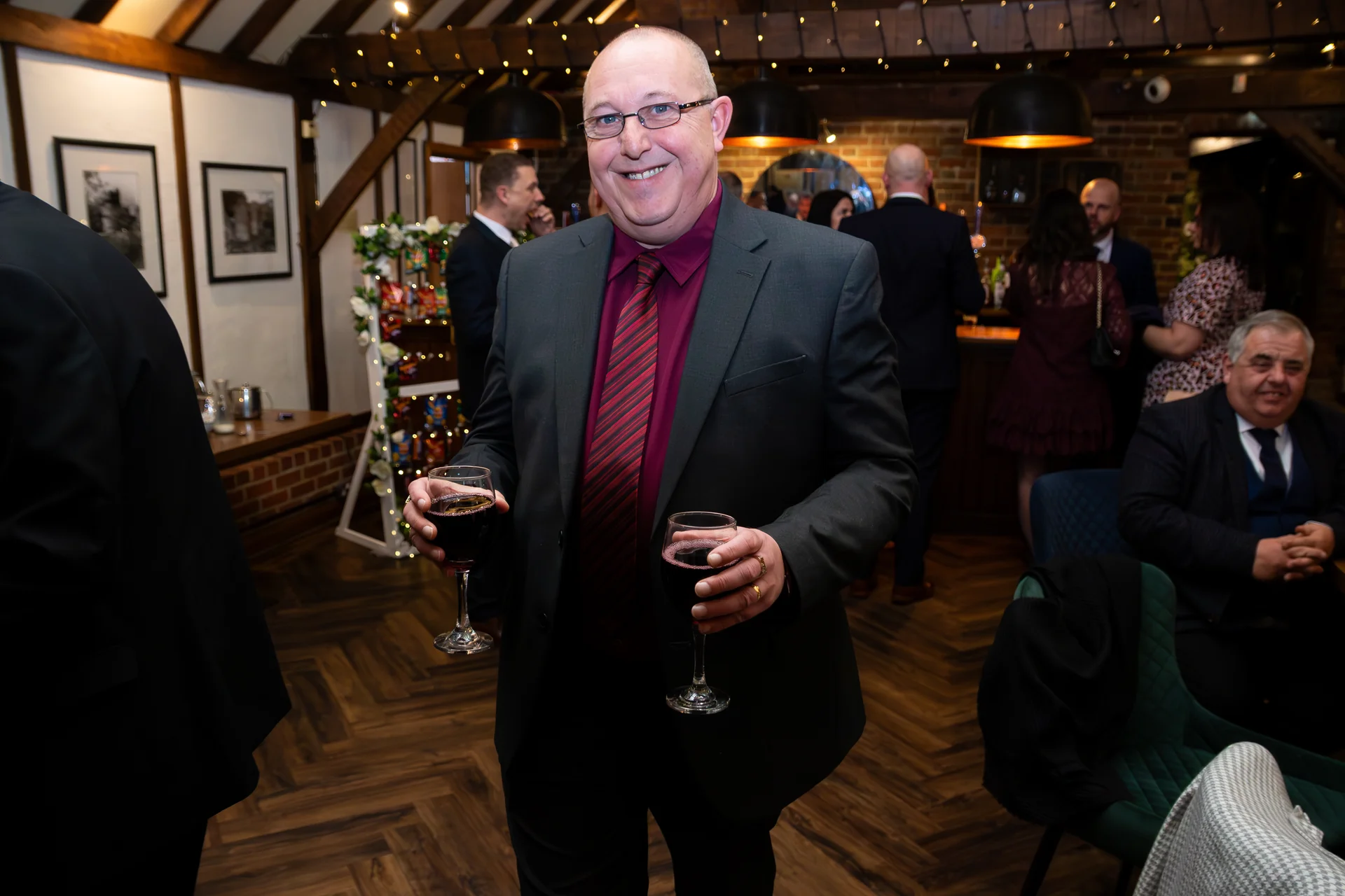 A smiling male wedding guest holding two glasses of red wine inside a rustic barn venue with exposed wooden beams and fairy lights.