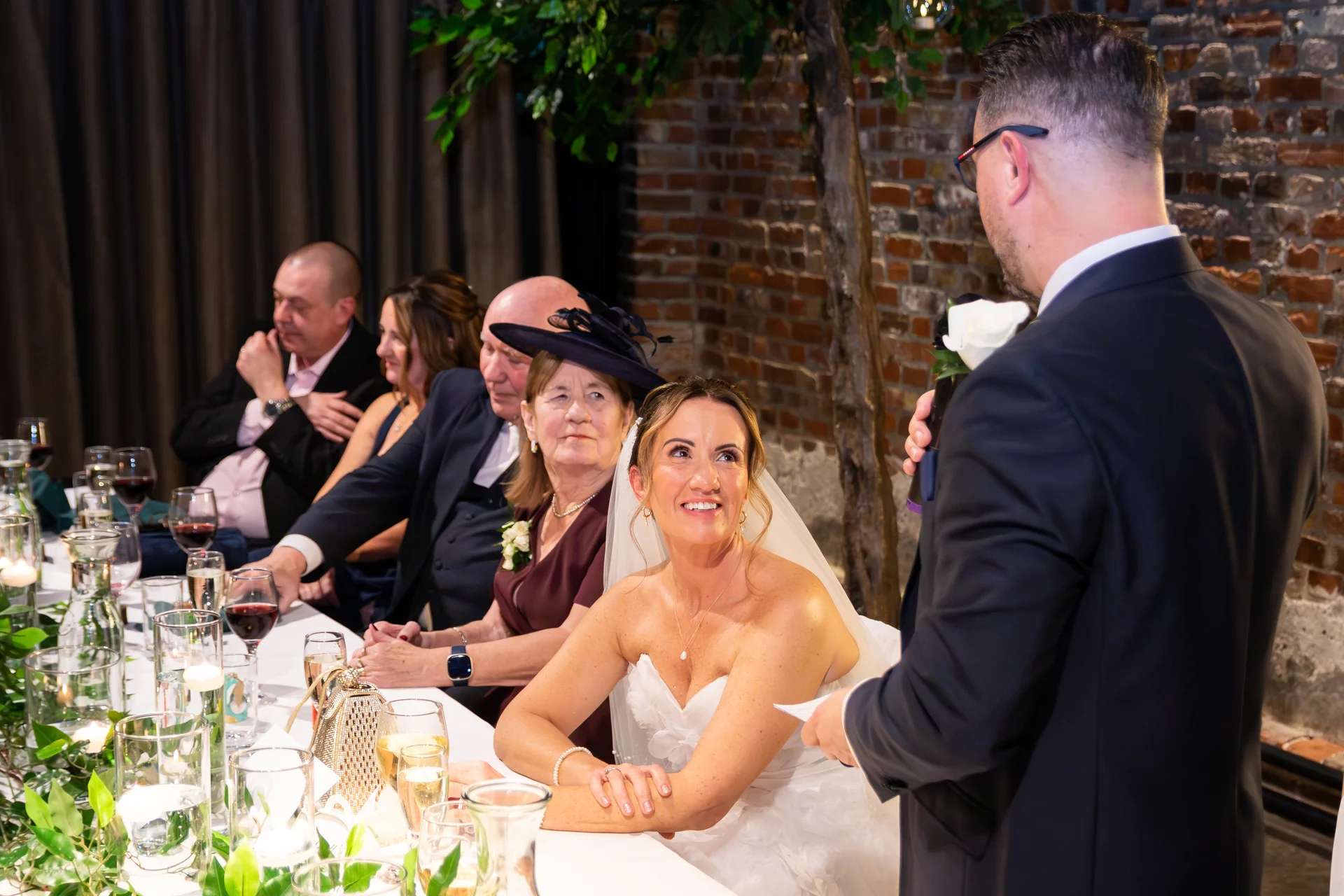 The bride smiles warmly while seated at the top table listening to a wedding speech, surrounded by guests in a reception venue with exposed brick walls.