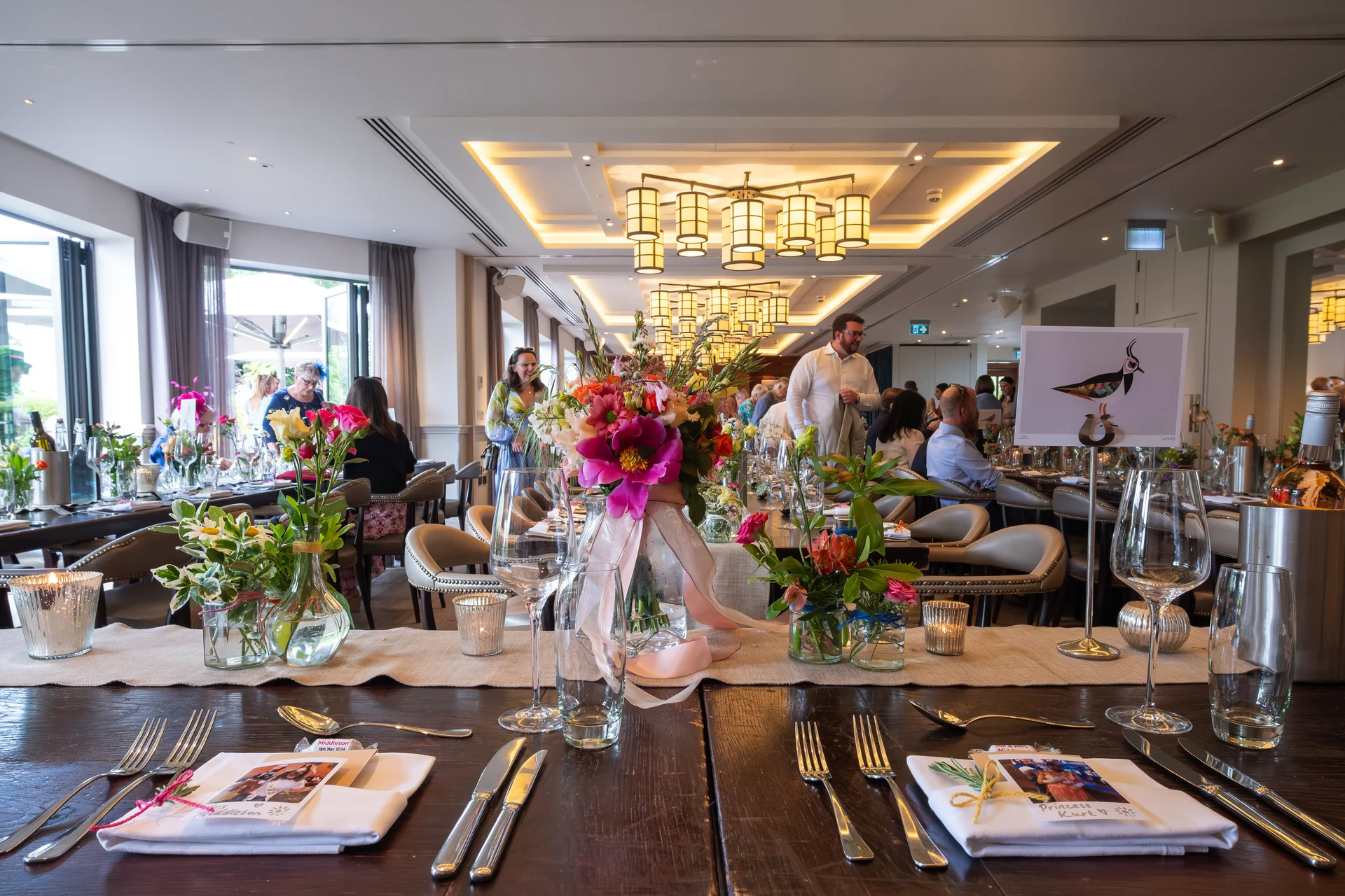 An elegantly styled long banquet table at a restaurant wedding reception features vibrant floral centerpieces, place settings with menus, wine glasses, and guests mingling in the brightly lit background.
