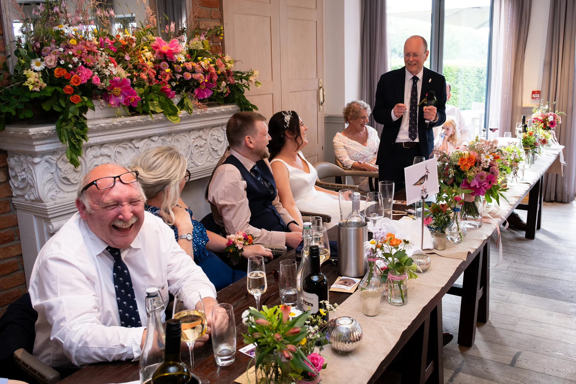 An older man stands and gives a speech at the top table during a wedding reception, while guests laugh joyfully in front of a decorated fireplace.