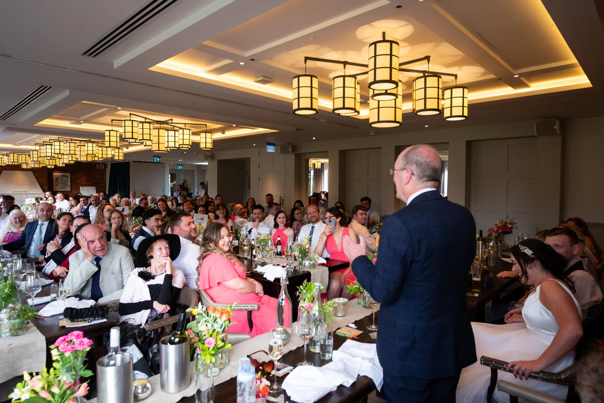 A guest delivers a speech at the top table while seated wedding guests watch, laugh, and listen during the wedding reception.