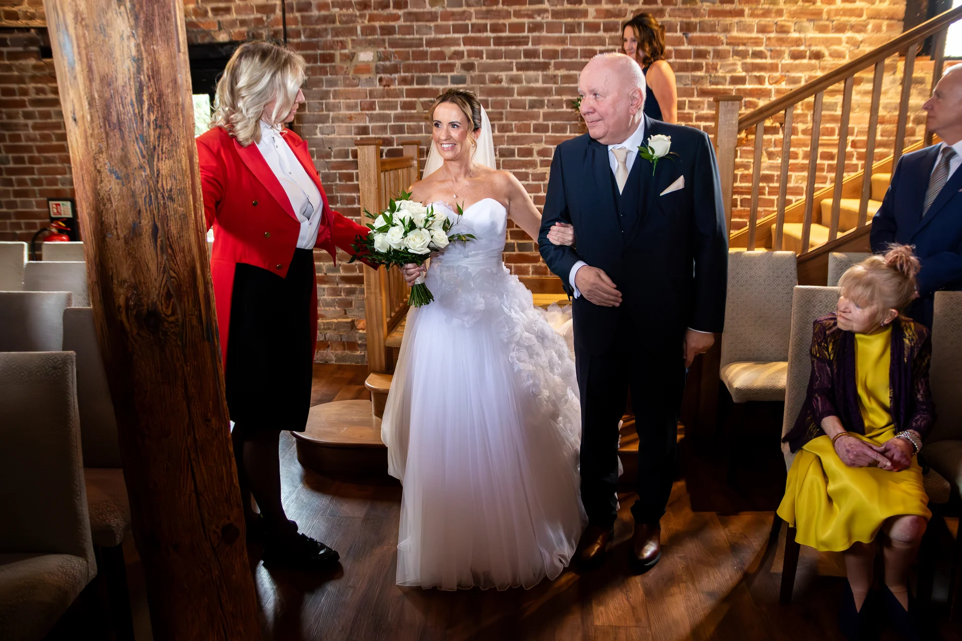 A smiling bride walks down the aisle arm in arm with her father inside a rustic barn venue with exposed brick walls.