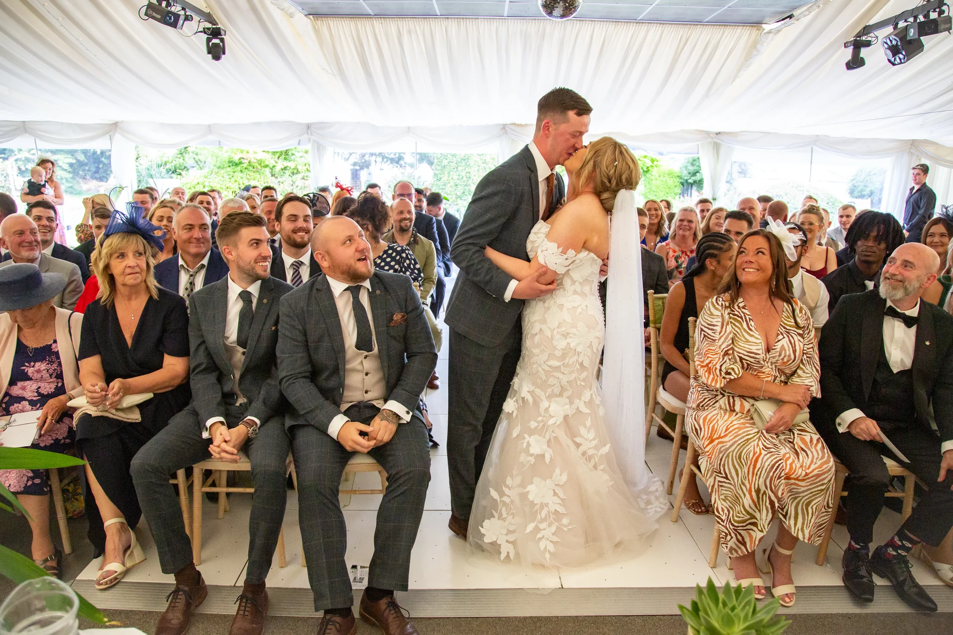 A bride and groom share their first kiss at the front of a marquee wedding ceremony, while seated guests look on and smile.