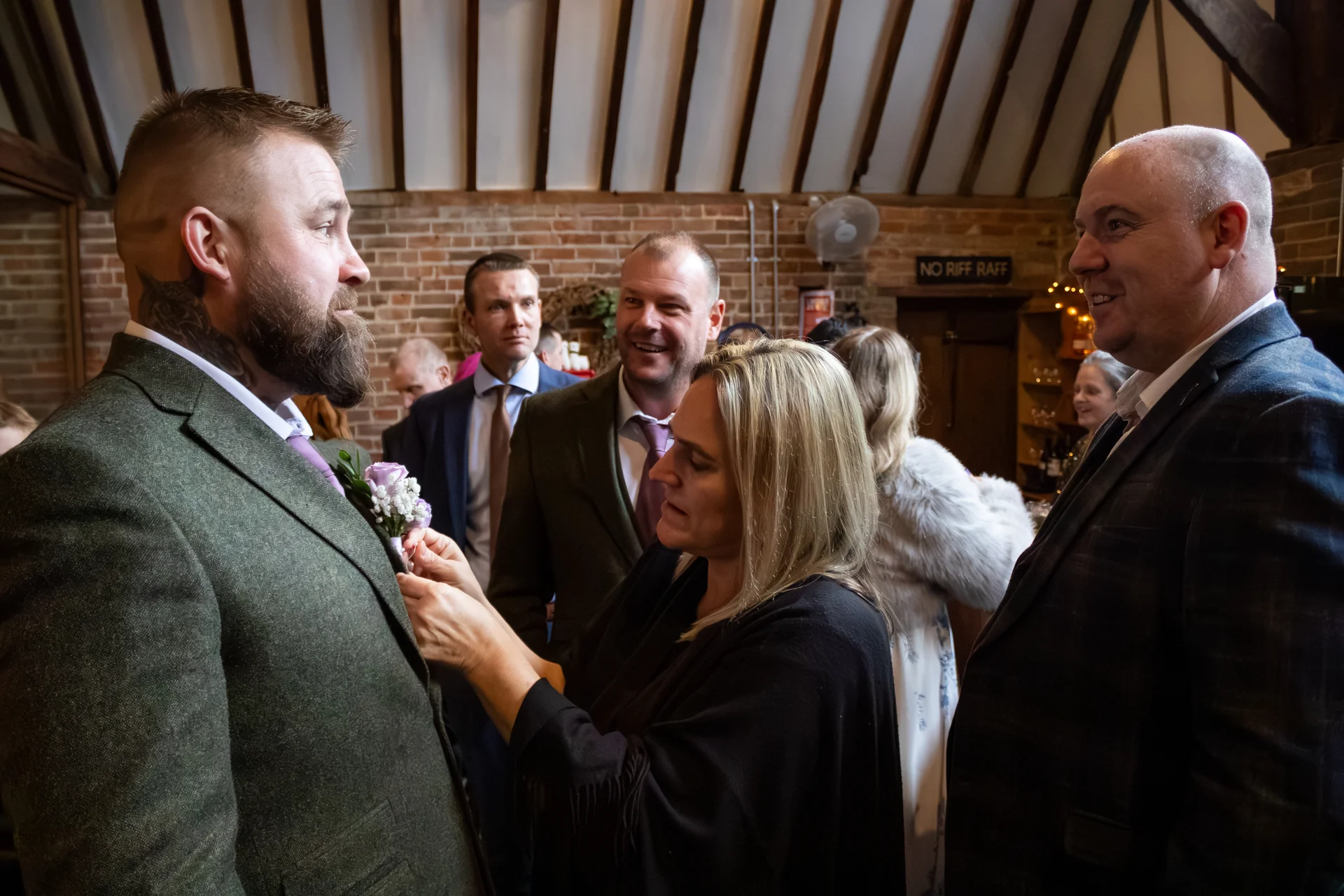 A guest carefully pins a floral boutonniere to the groom's green tweed jacket inside a rustic barn venue with exposed brick walls.