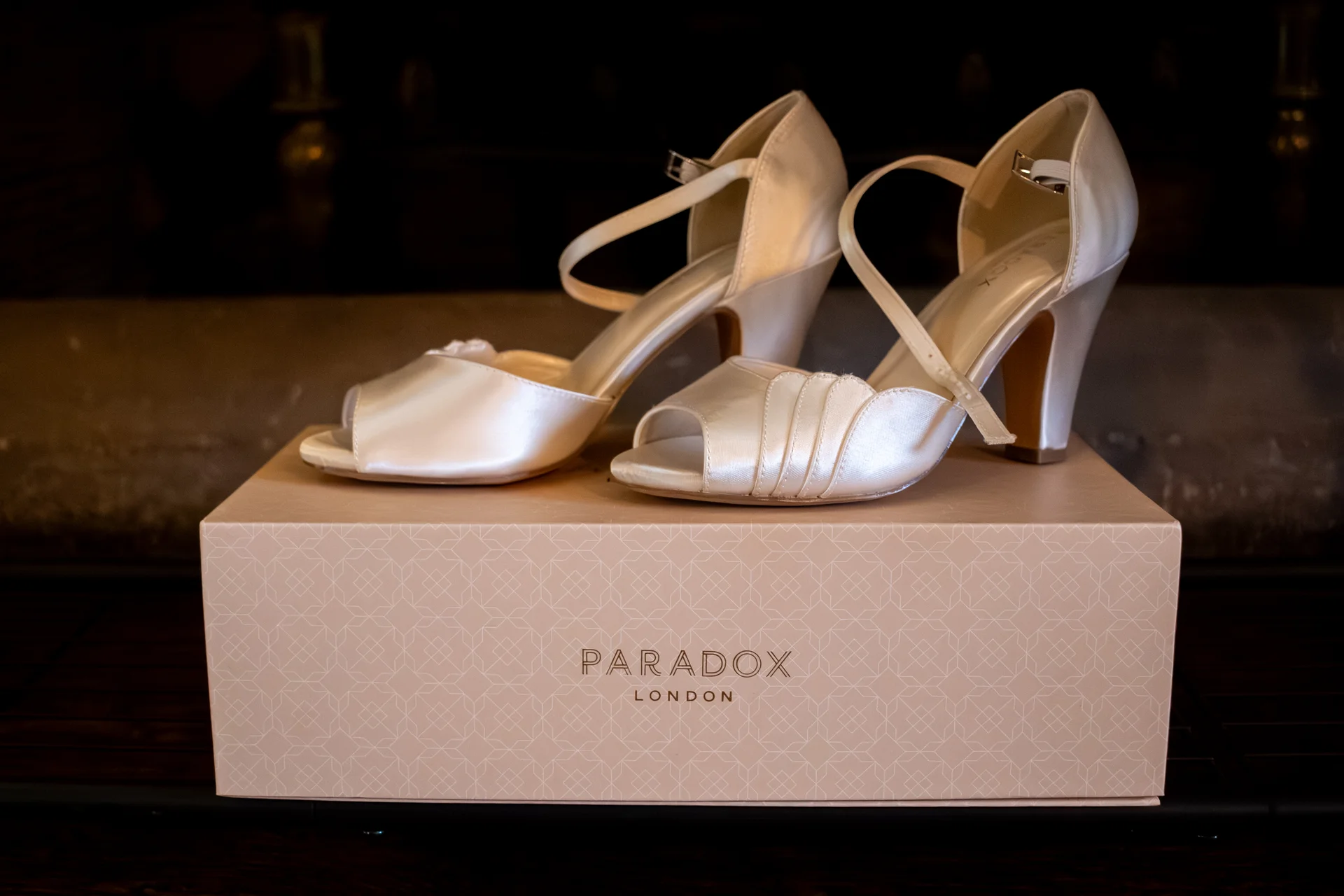 A pair of ivory satin wedding heels from Paradox London resting elegantly on top of their branded shoebox during bridal preparations.