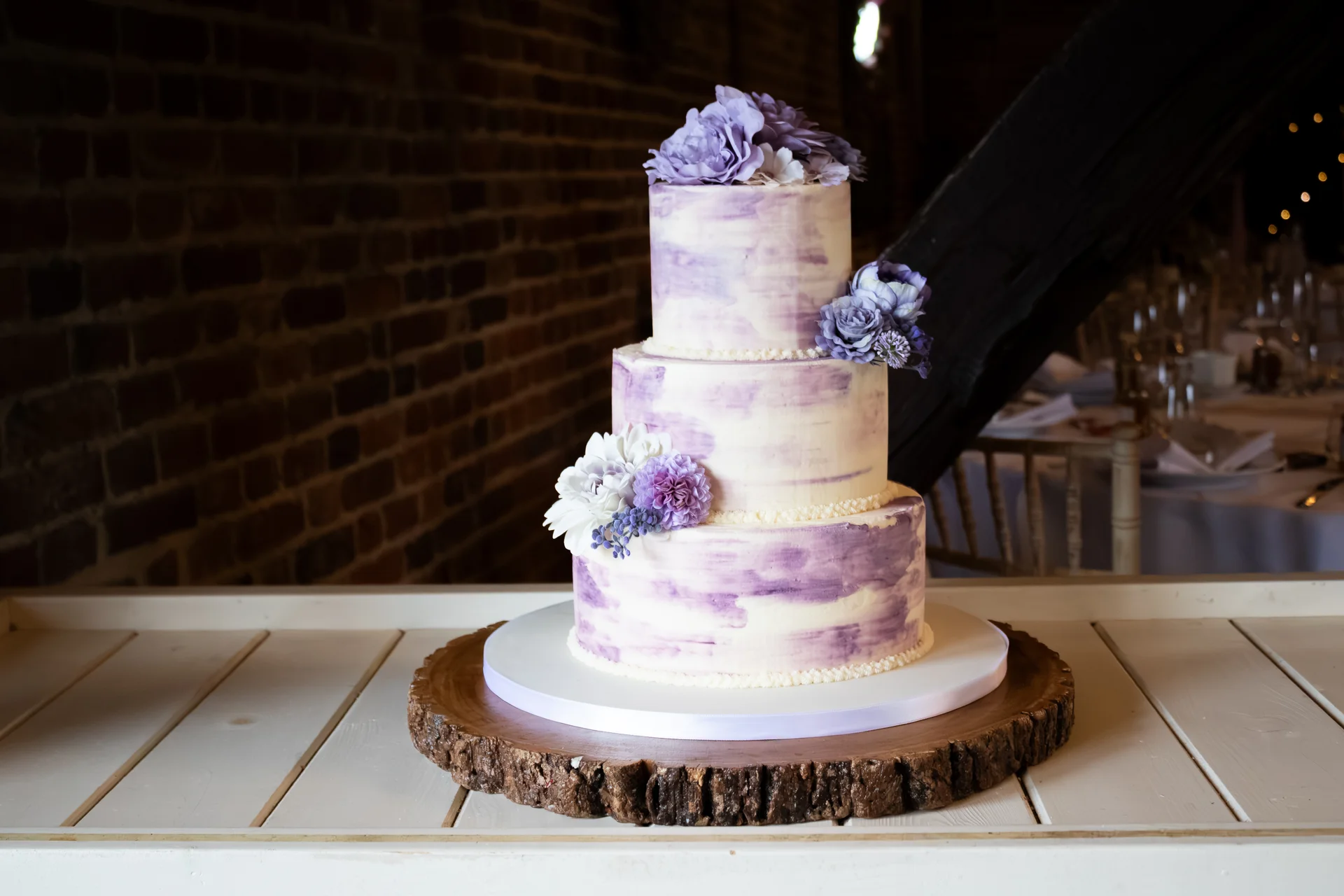 A three-tier watercolour buttercream wedding cake with purple and white floral decorations sits on a rustic wood slice stand against an exposed brick wall.