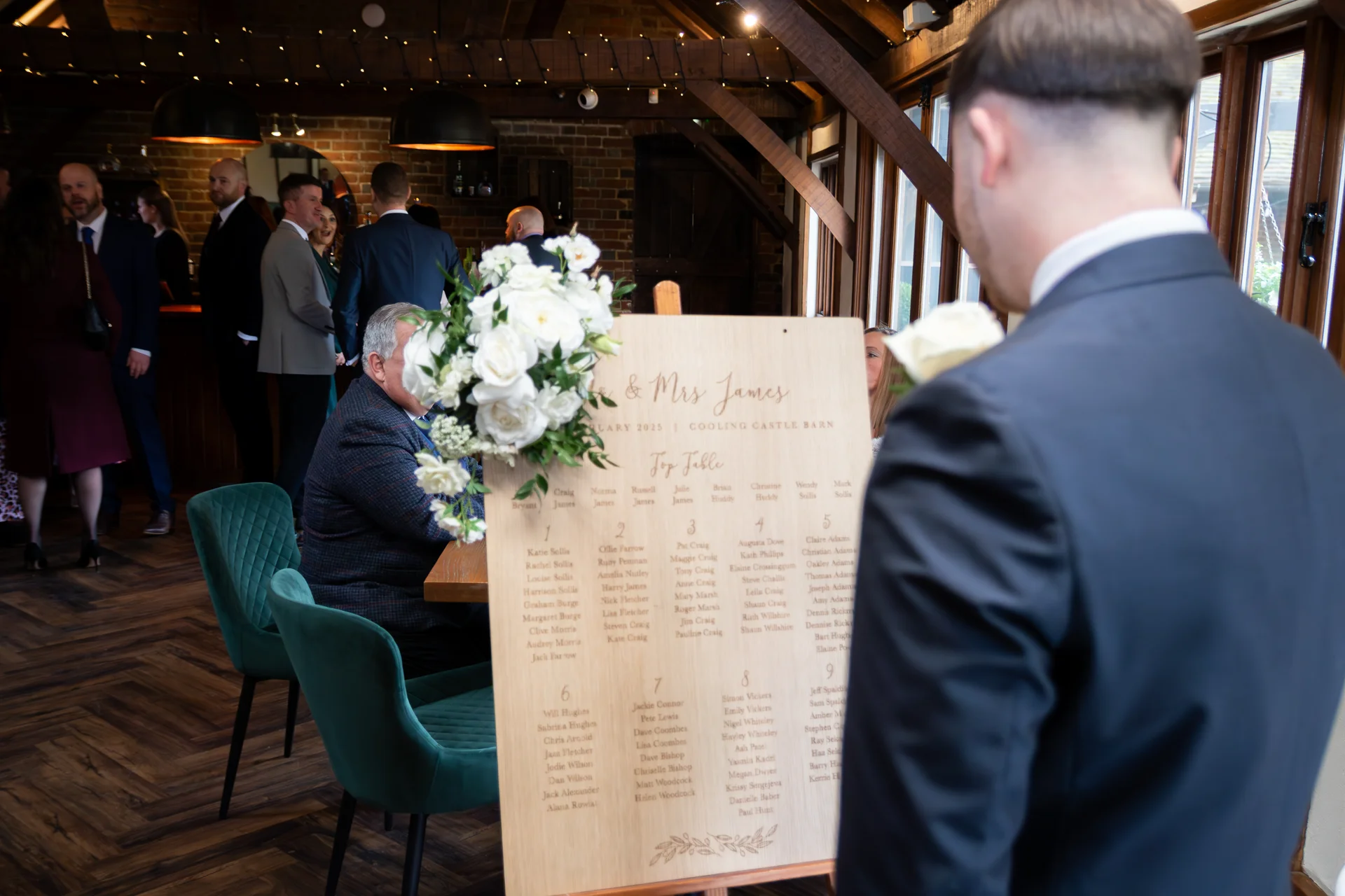 An over-the-shoulder view of a guest in a dark suit looking at a wooden wedding seating plan adorned with white roses inside a rustic barn venue.