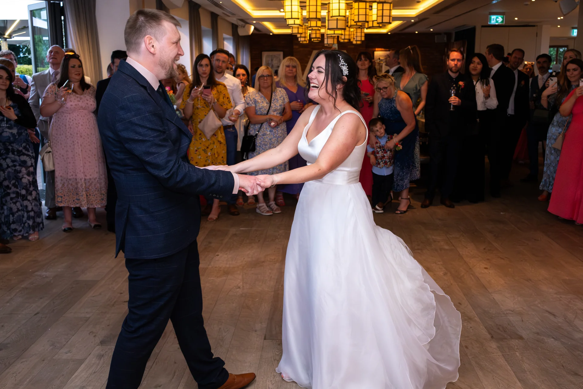The bride and groom share a joyful first dance on a wooden floor, surrounded by smiling wedding guests inside a well-lit reception room.