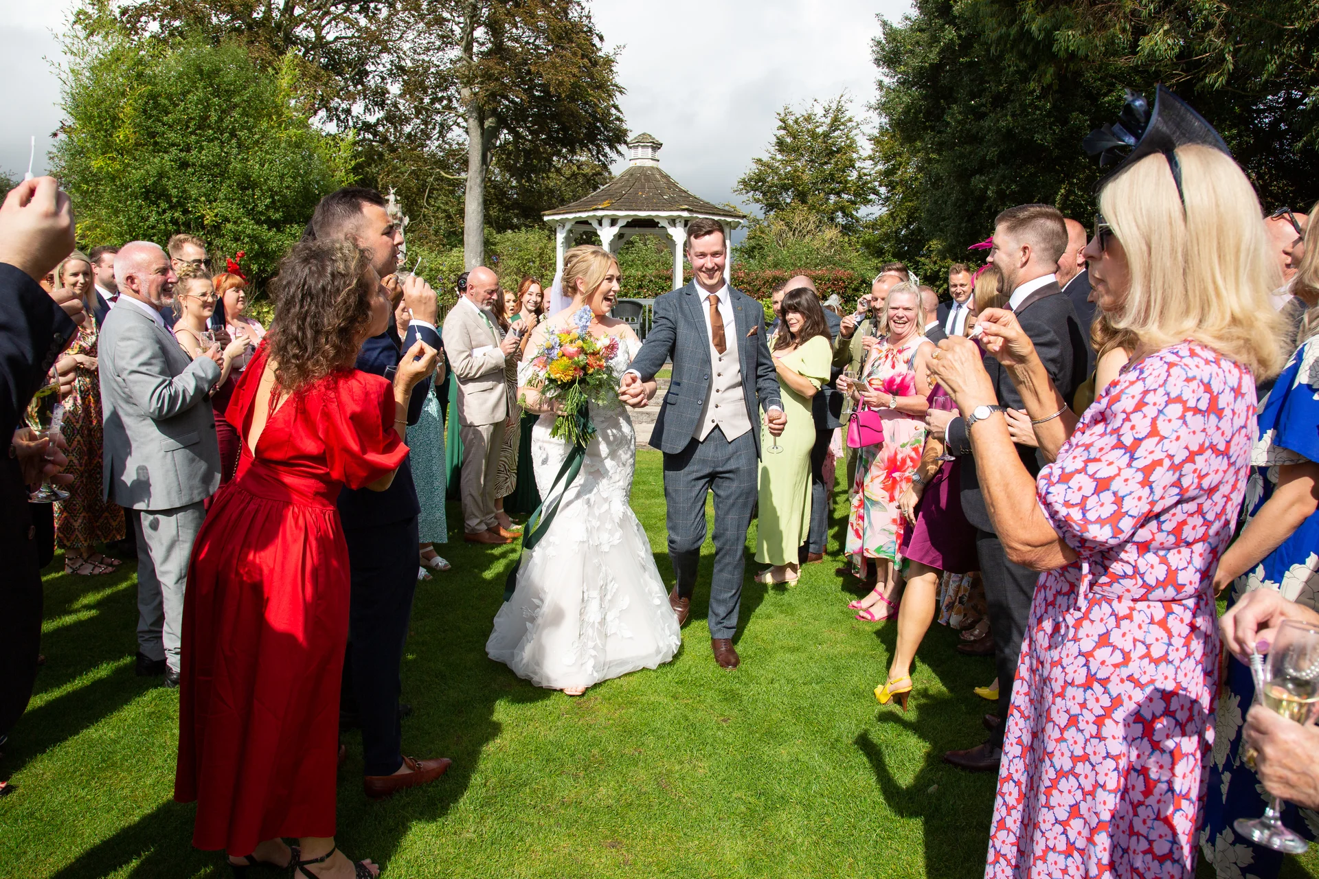 The smiling bride and groom walk hand in hand through an outdoor garden reception as cheering guests blow bubbles around them.