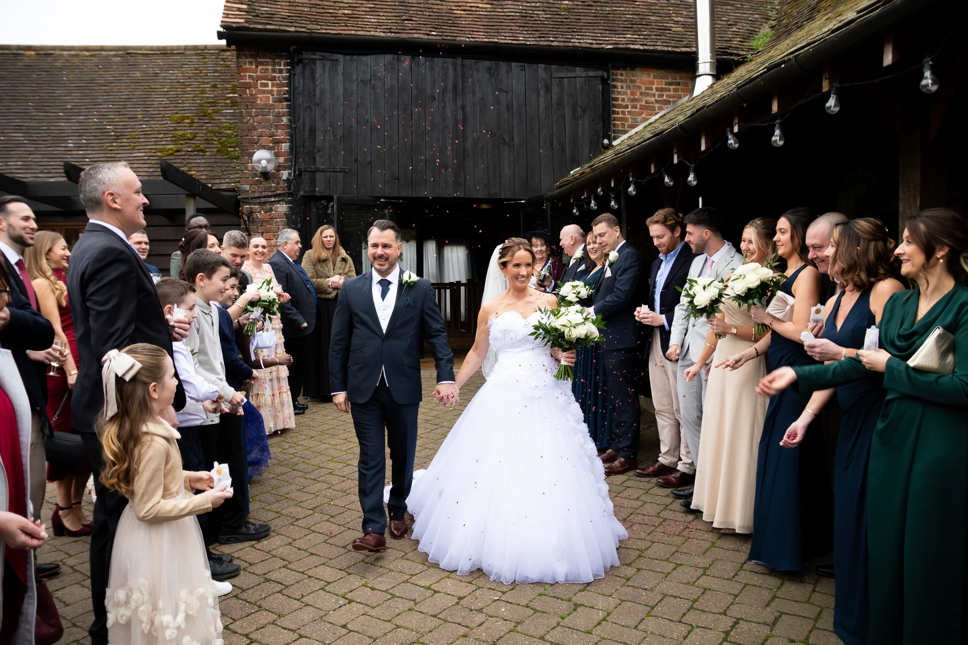 Newlyweds walk hand in hand through a joyful crowd of guests throwing confetti in a rustic barn courtyard.
