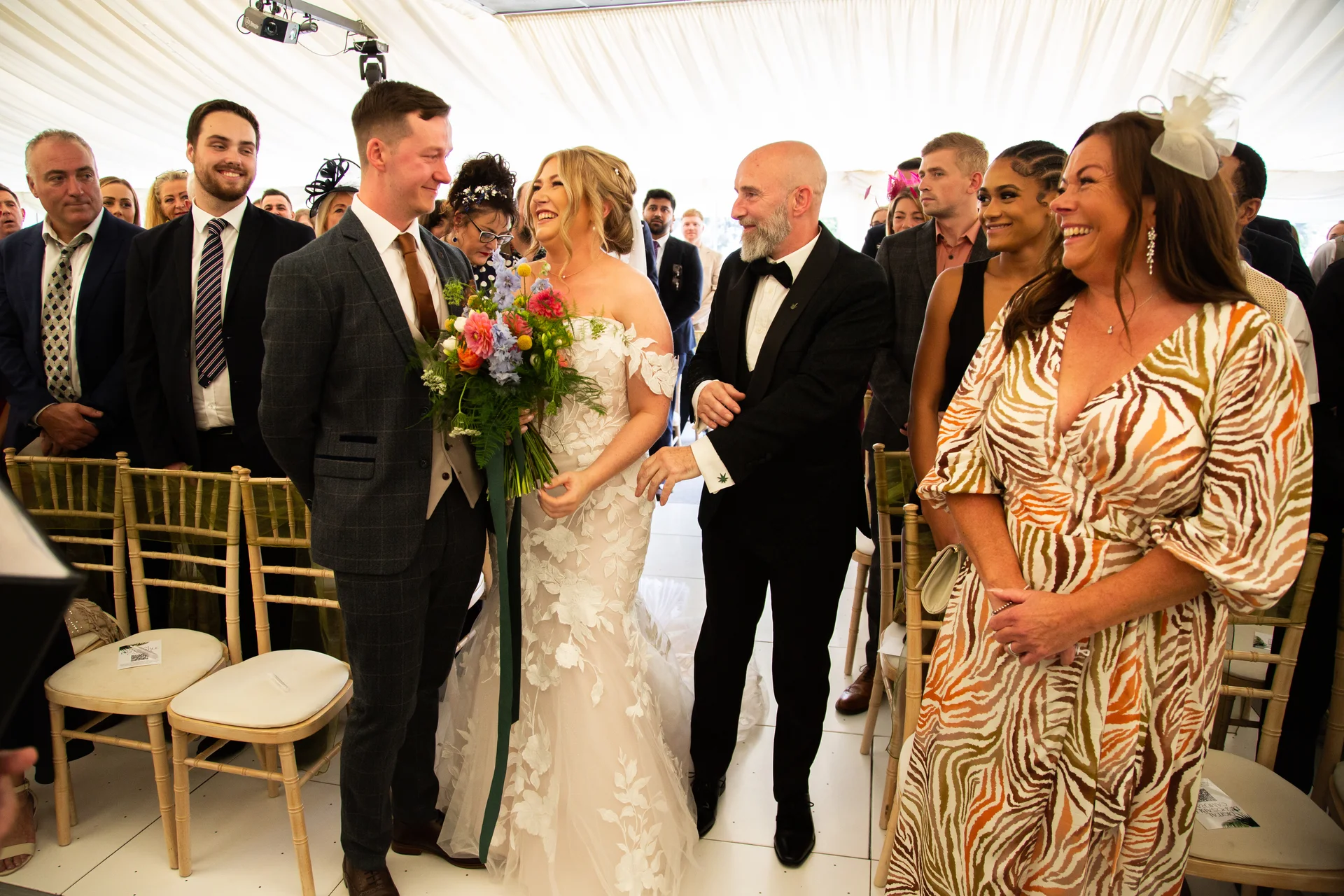 A happy bride and groom stand together holding a colourful bouquet while guests look on and smile inside a bright marquee wedding venue.