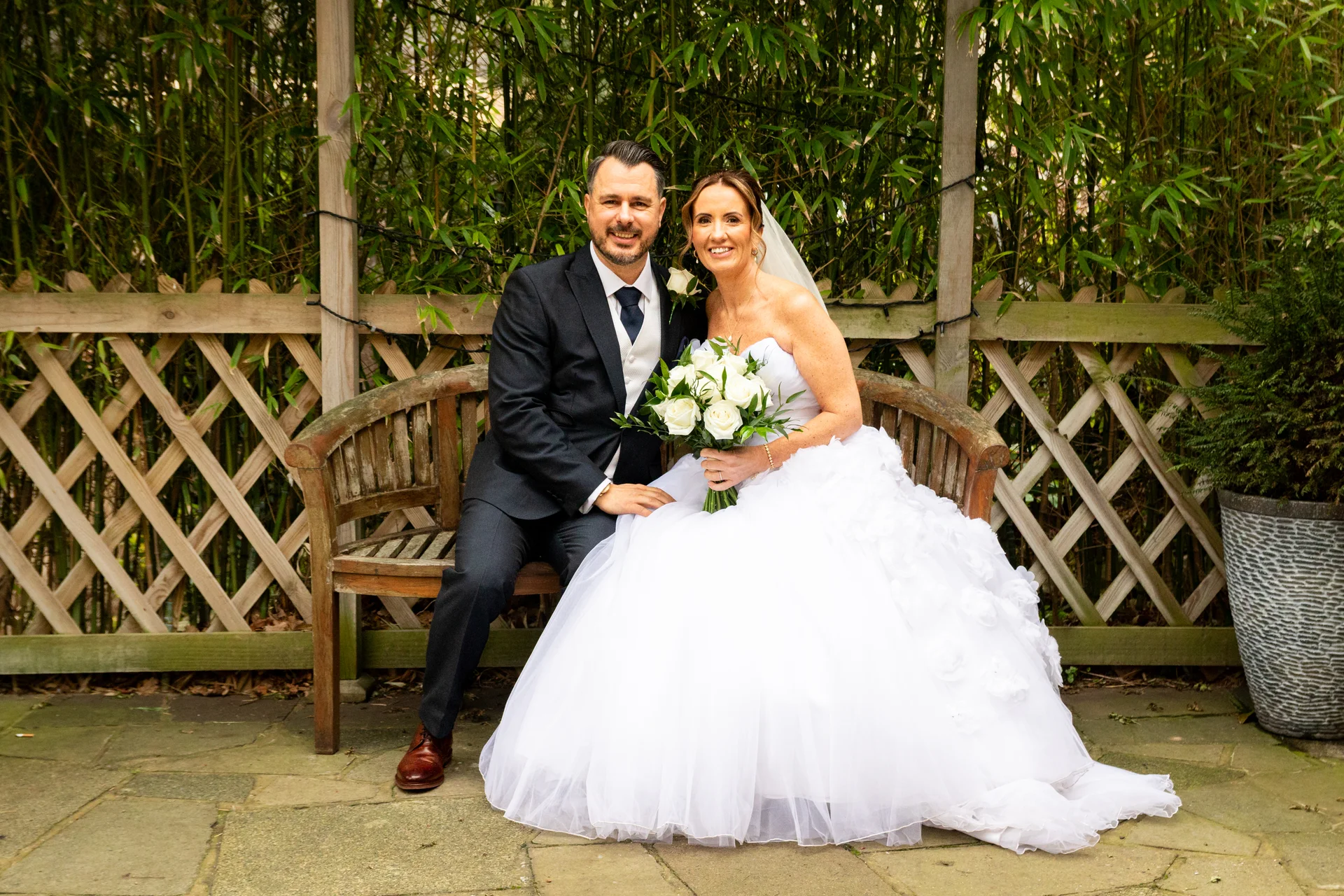 A newly married couple sit together on a wooden garden bench, the bride holding a bouquet of white roses while wearing a strapless ball gown.