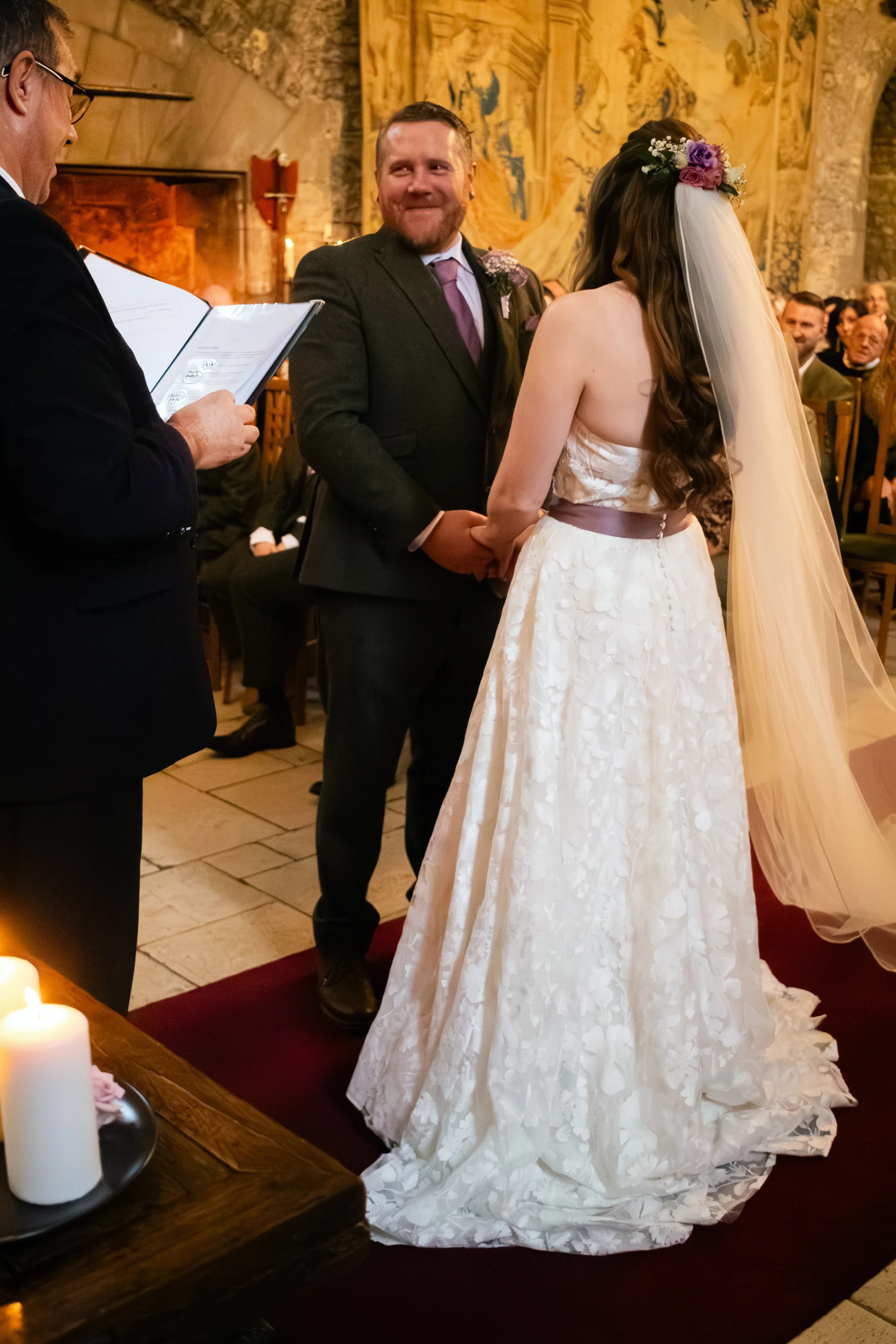 The bride and groom stand holding hands during their civil ceremony inside a historic stone castle venue while guests watch from the pews.