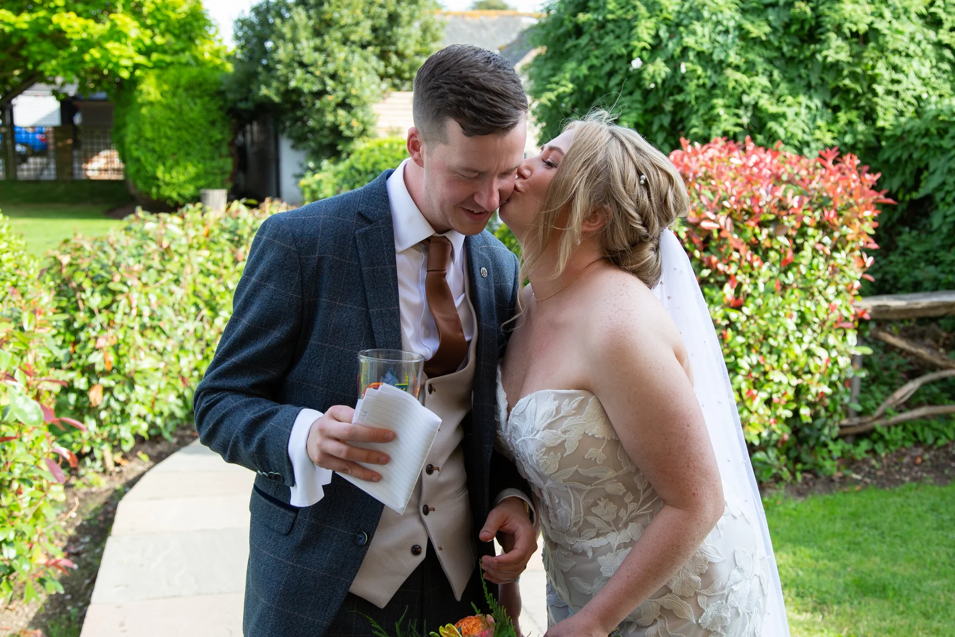 The bride kisses the groom on the cheek outdoors in a lush garden setting during their summer wedding celebration.